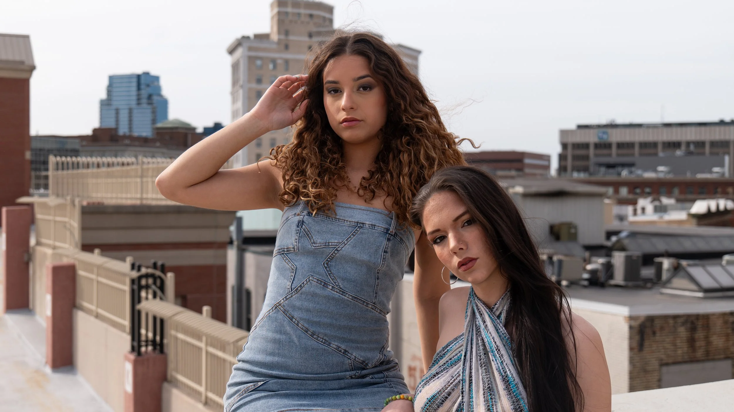 Two young women with long dark hair pose on a rooftop against a city skyline, one with curly hair in a denim dress and the other with straight hair in a patterned top.