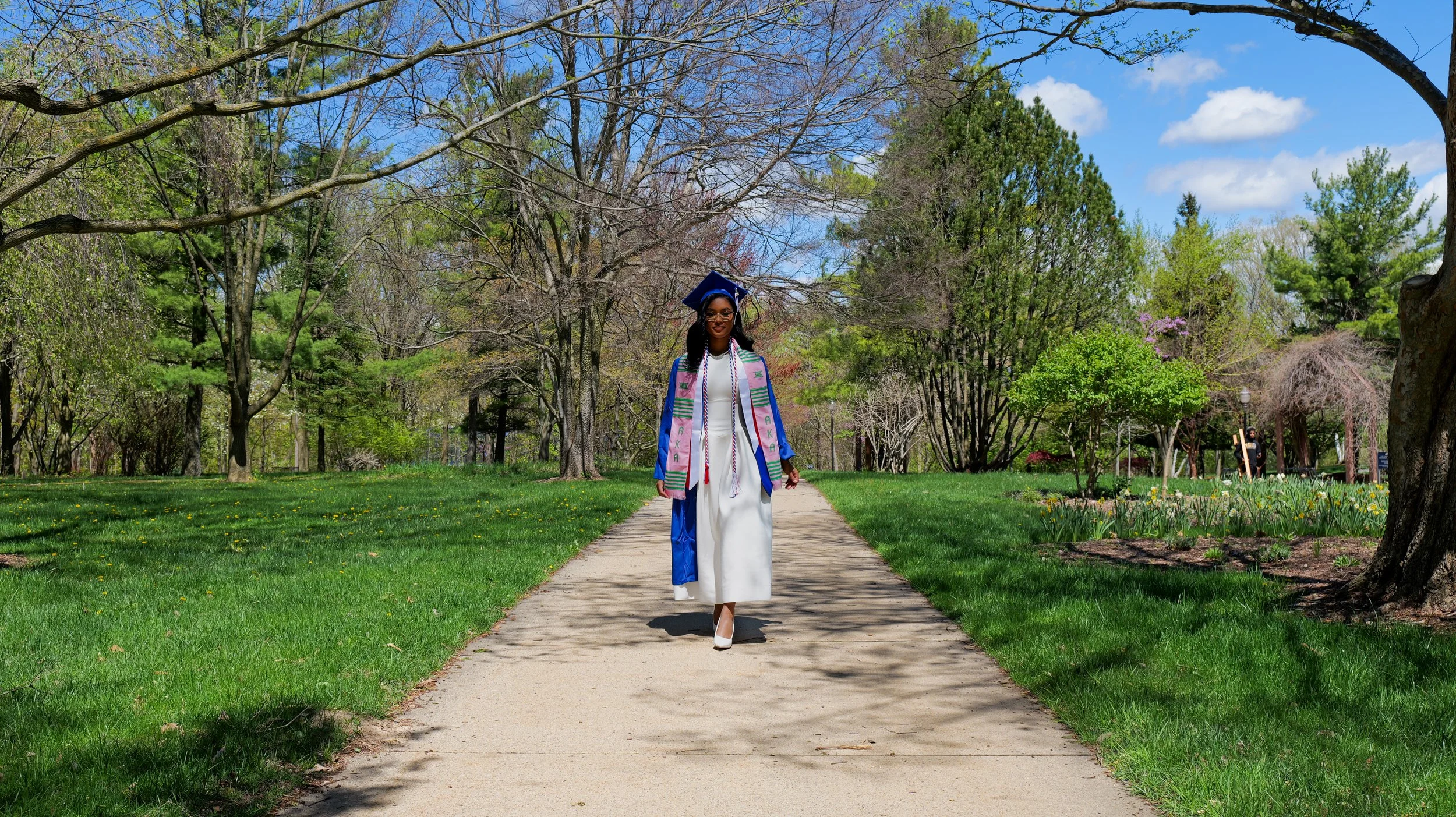 A woman in a white dress and blue graduation gown walking on a park pathway, wearing sunglasses and a graduation cap.