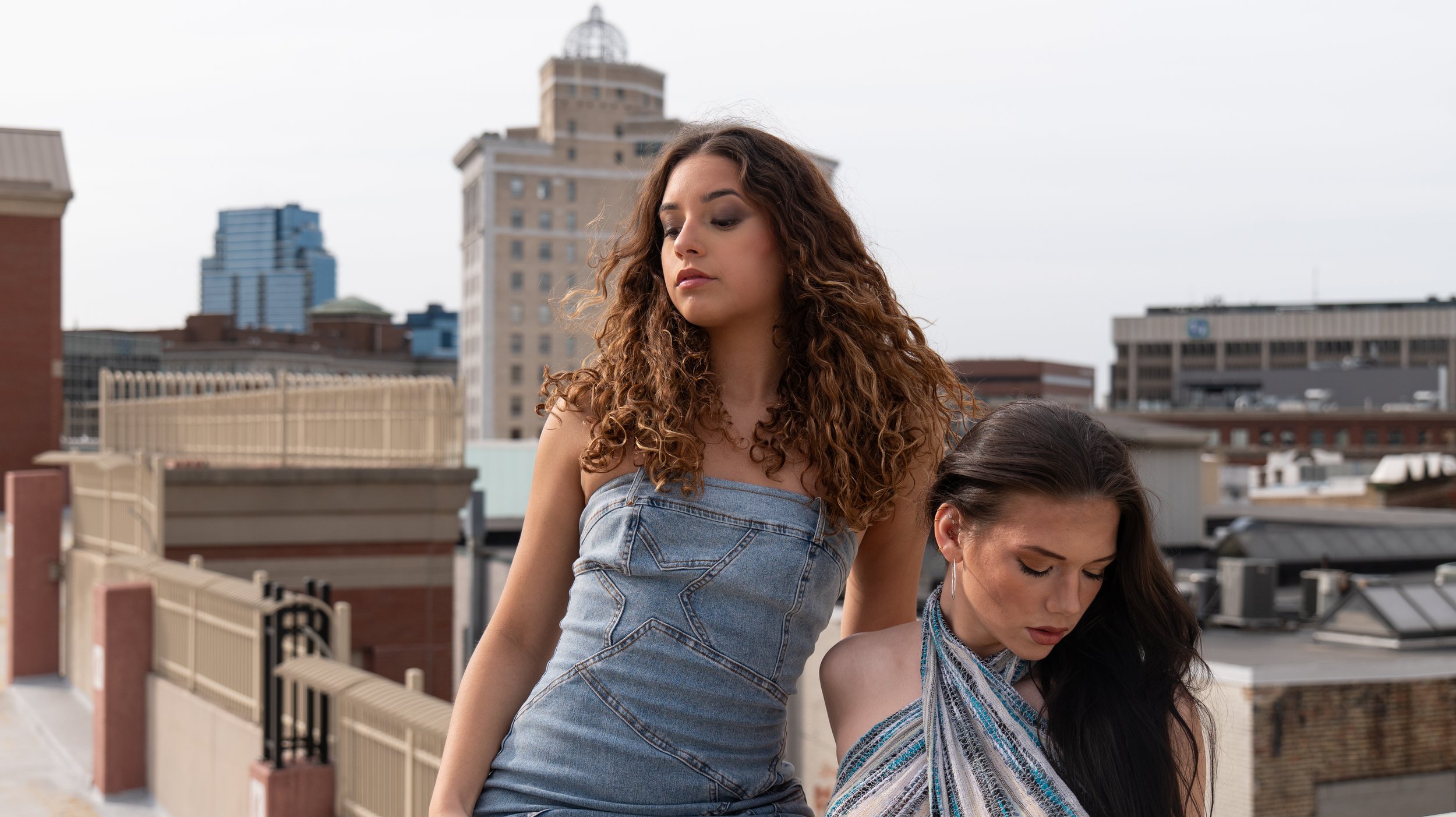 Two women standing on a rooftop with city skyline in the background, one with curly brown hair looking down, the other with dark straight hair looking down.