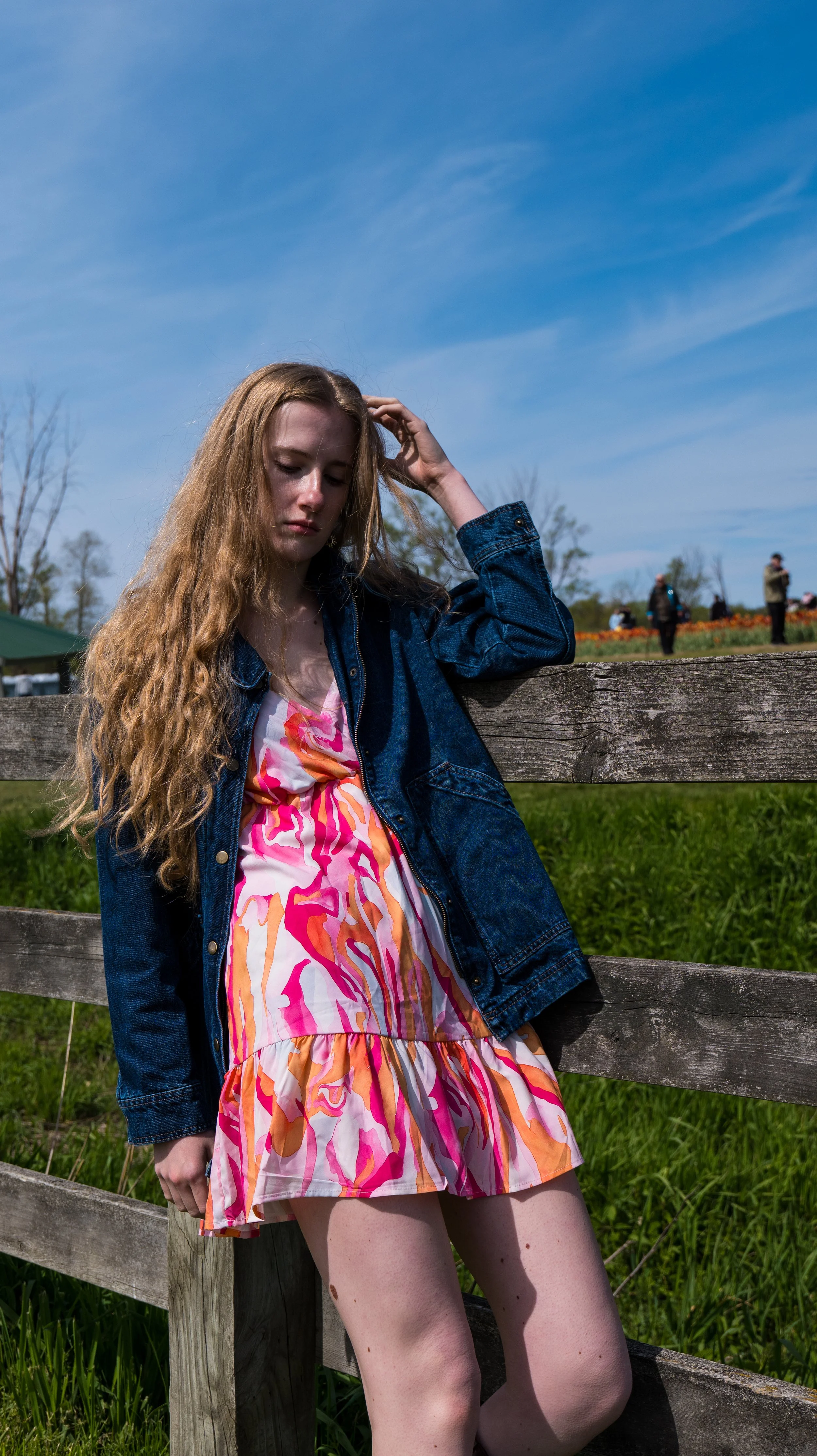 A young woman with long curly hair leaning against a wooden fence outdoors. She is wearing a colorful dress with pink, orange, and white patterns and a denim jacket. In the background, there are people and trees under a clear blue sky.