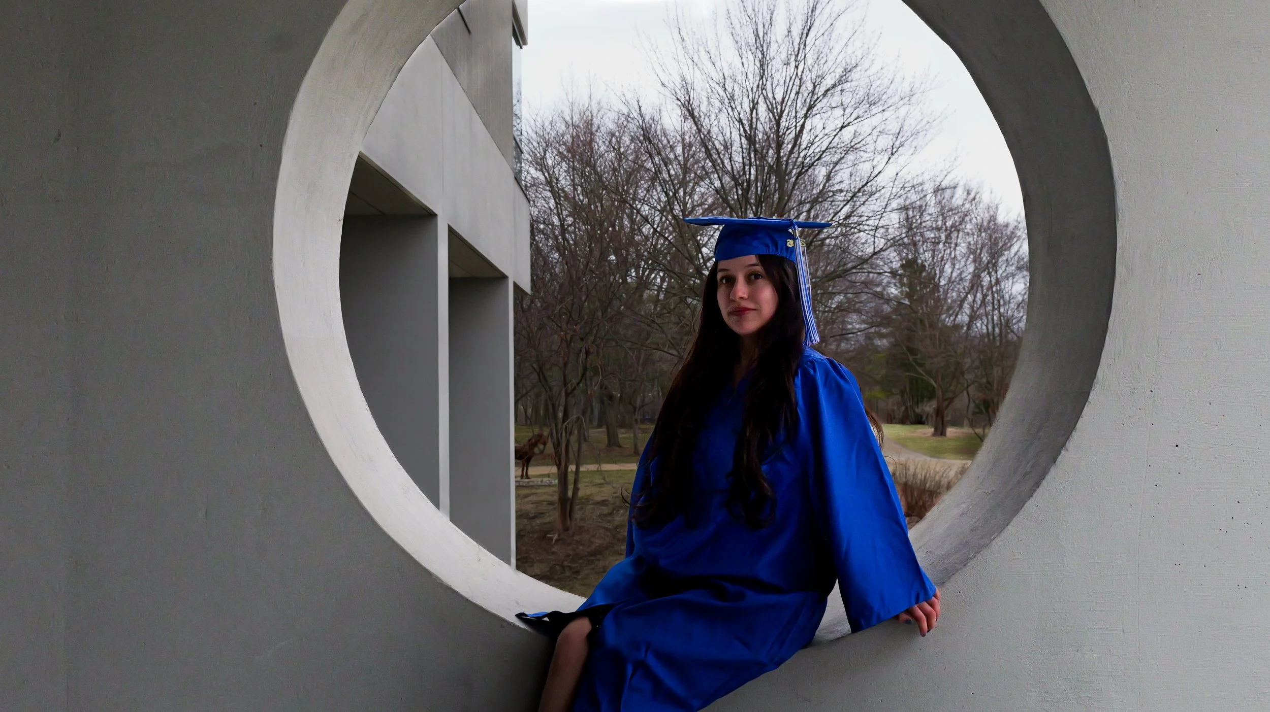 Young woman in a blue graduation gown and cap sitting and posing inside a circular architectural structure with trees and a park in the background.