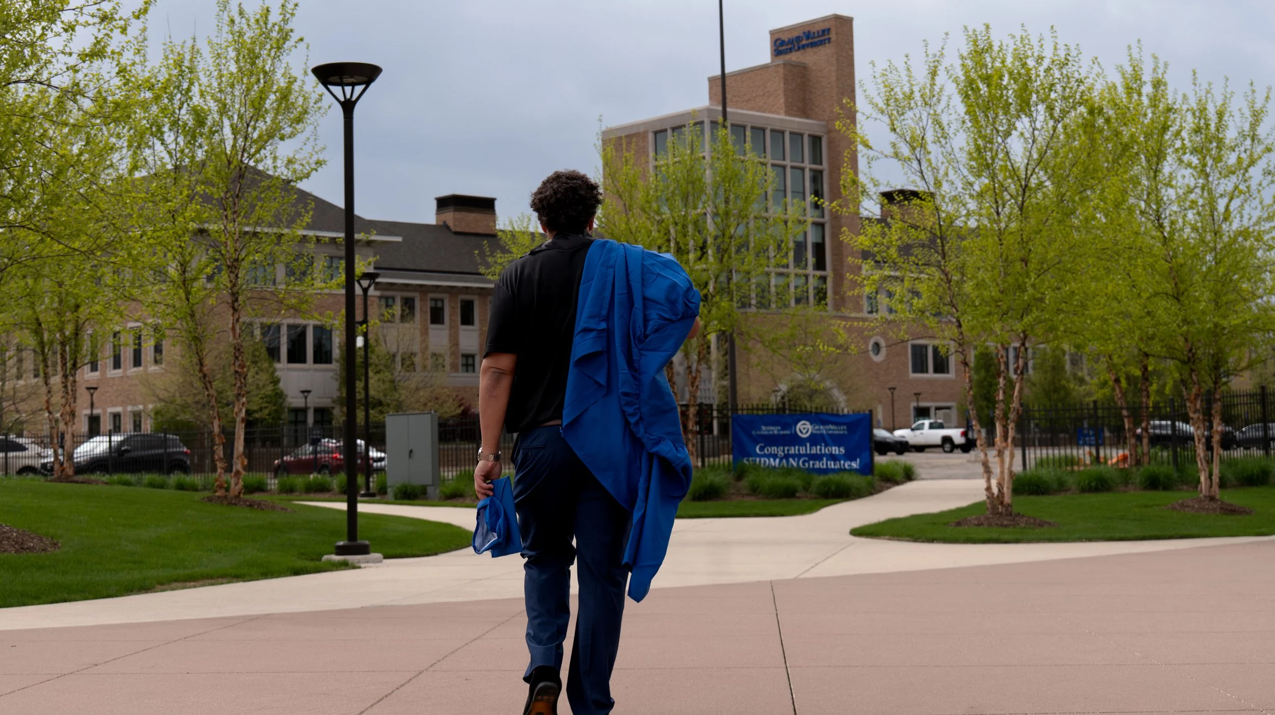 A person walking on a paved pathway in a college campus courtyard, carrying a blue jacket over their shoulder, with a building and trees in the background.