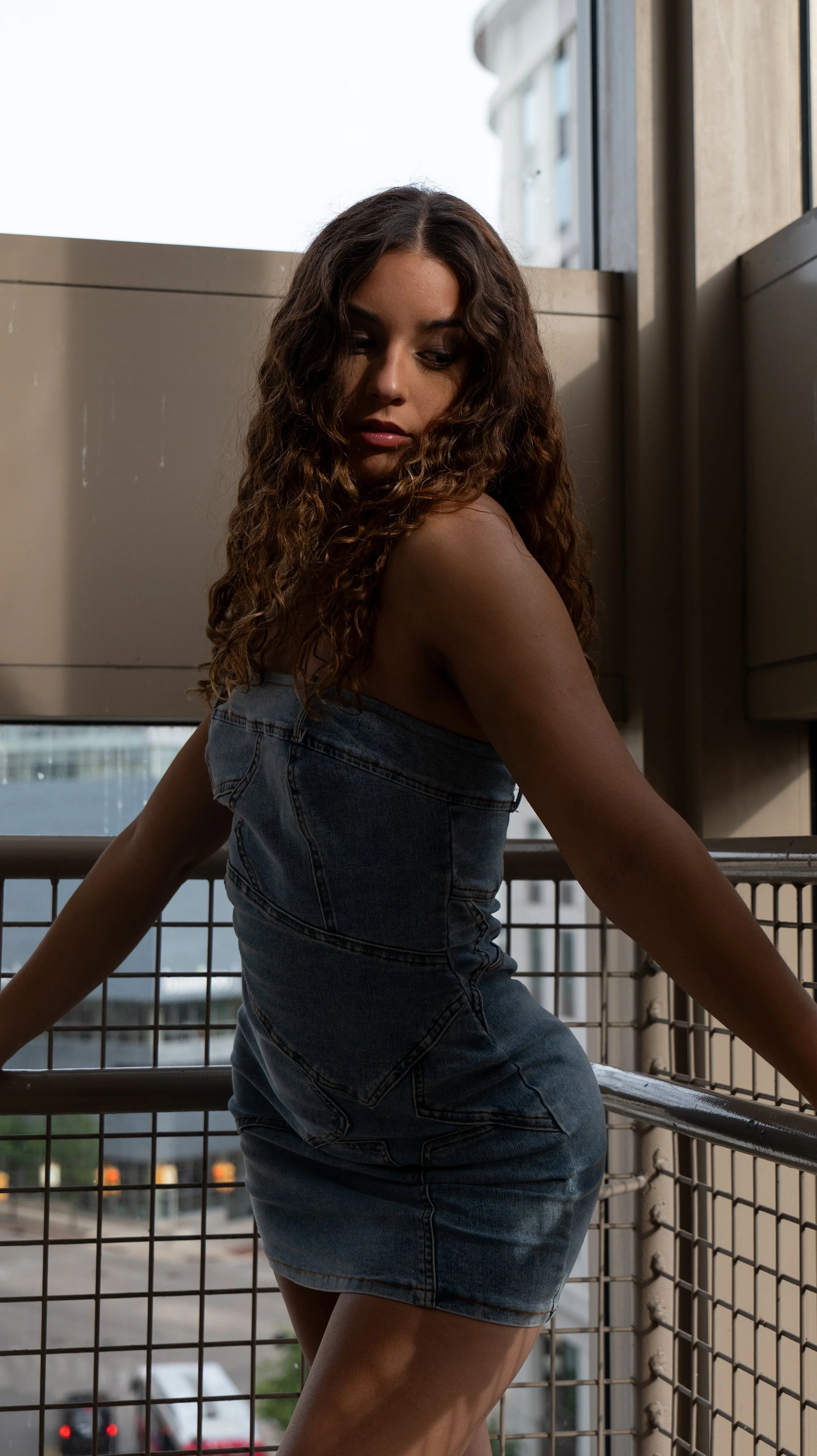 A young woman with long curly hair in a denim dress is sitting on a balcony railing beside a metal fence, looking downward with a thoughtful expression, behind a cityscape with tall buildings and a partly cloudy sky.