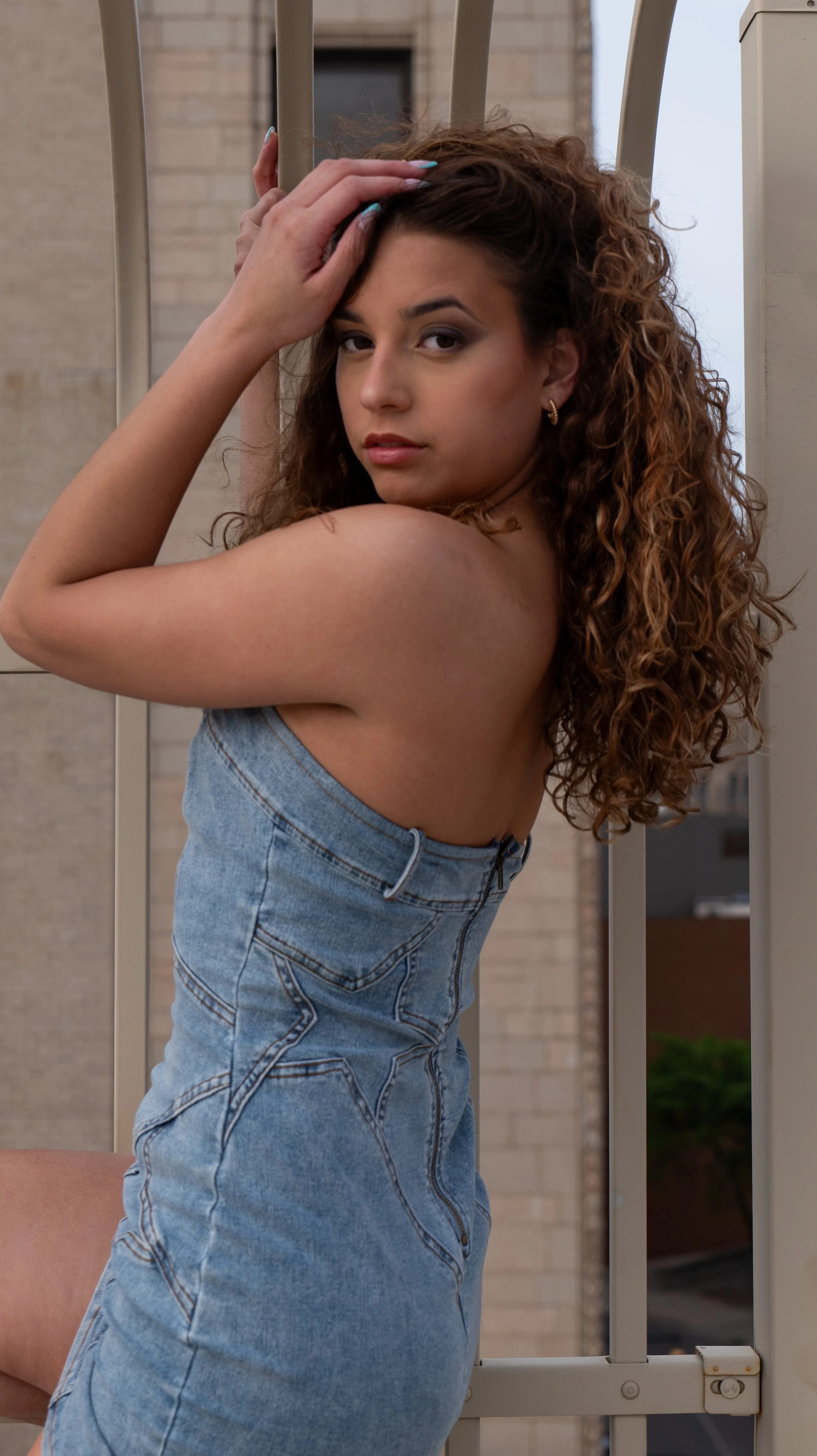 Young woman with curly hair wearing a denim dress, leaning on a metal railing, looking at the camera with a neutral expression.