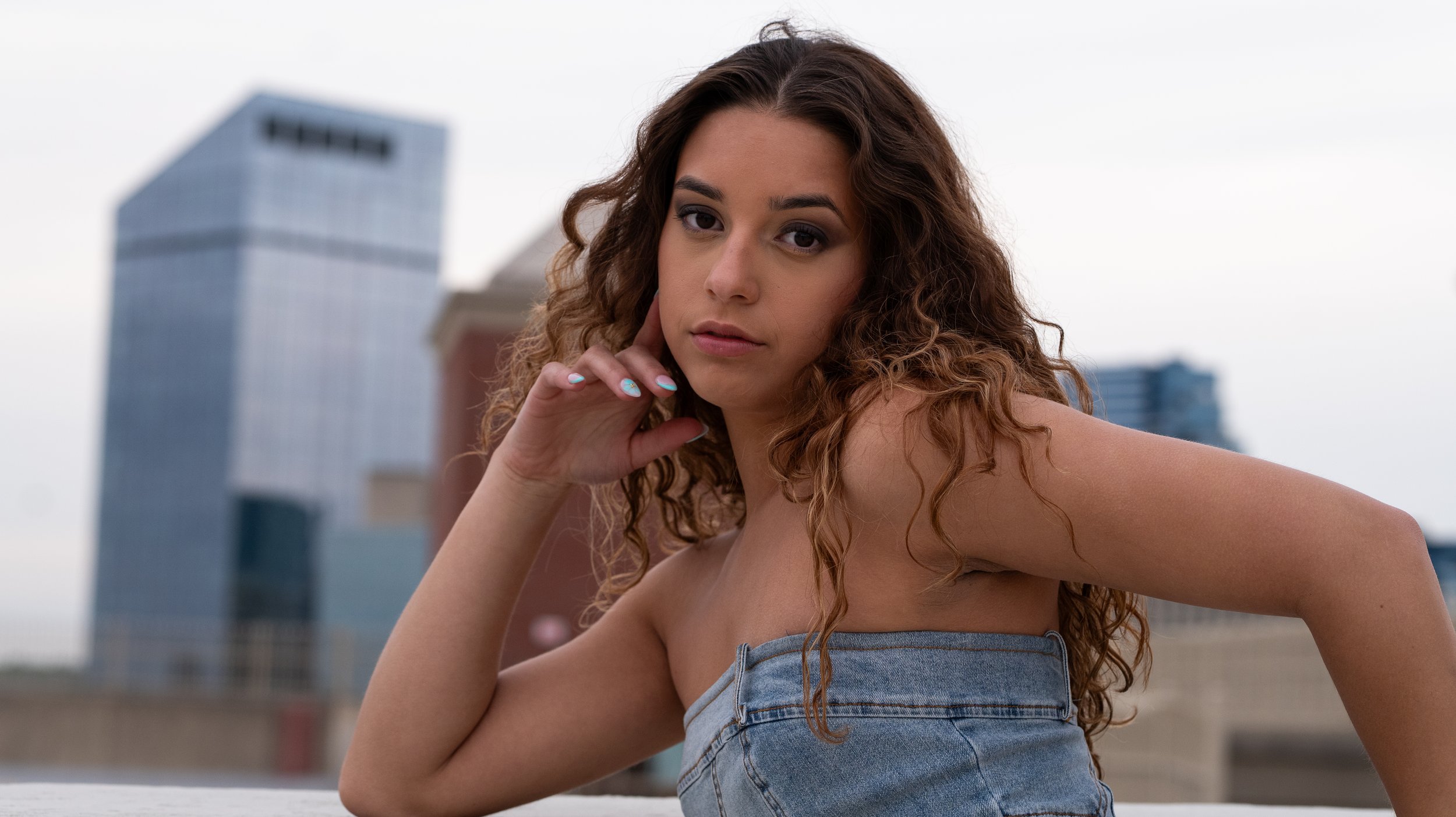 A young woman with curly hair and a strapless denim top posing outdoors in front of a cityscape with modern buildings.