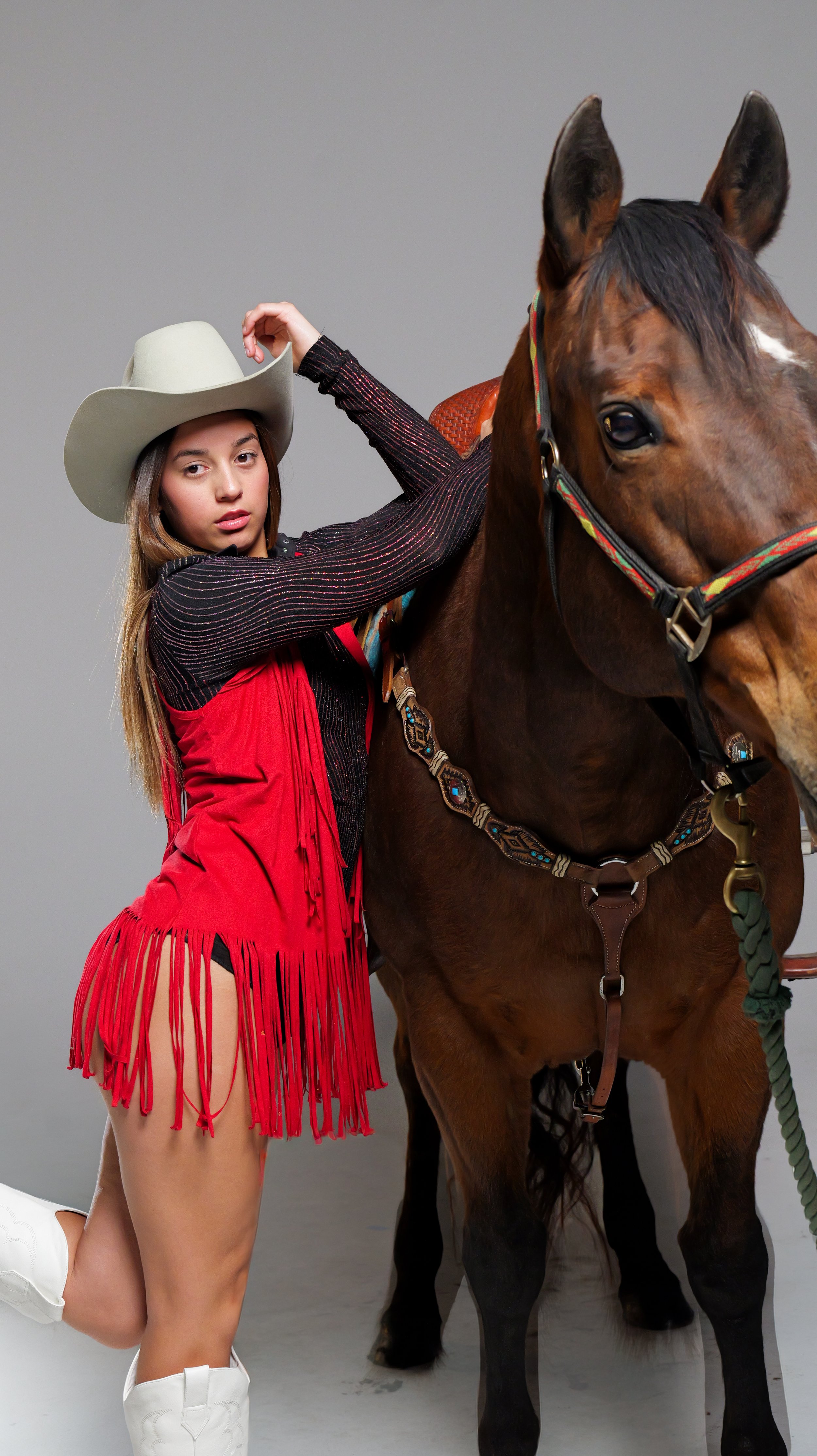 A young woman wearing a large white cowboy hat, a black long-sleeve top with red and gold stripes, a red fringe dress, and white cowboy boots, standing next to a brown horse with a colorful bridle and saddle, against a plain gray background.