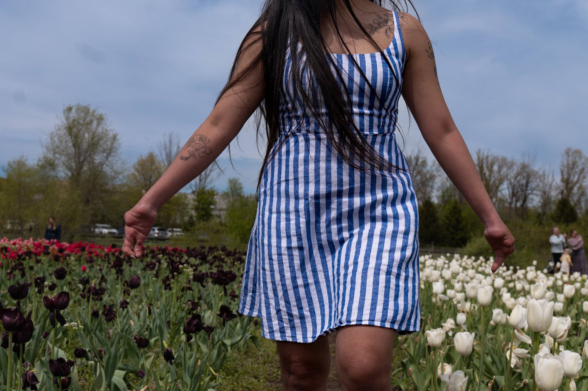 A woman in a blue and white striped dress standing in a tulip field with her arms outstretched, with trees and cloudy sky in the background.