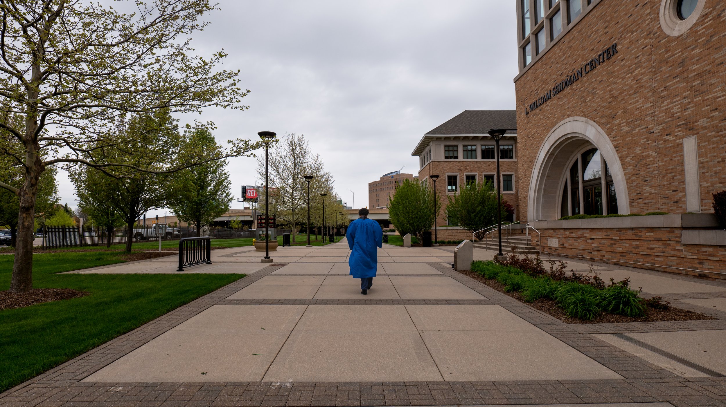 A person in a blue coat walking on a paved sidewalk outside William Sedman Center on a cloudy day, surrounded by grass and trees.