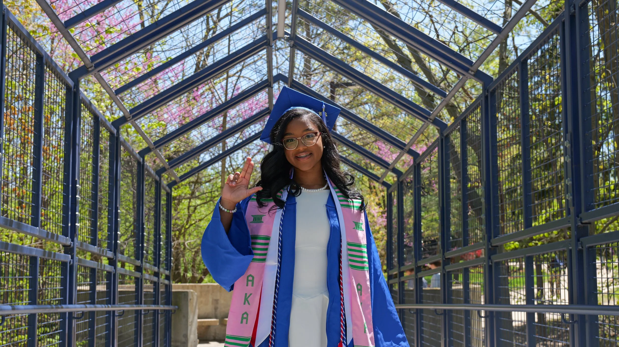A young woman in a blue graduation gown and cap, wearing glasses and a pink stole with green lettering, smiling and making a hand gesture, standing in a metal walkway with trees and pink flowers in the background.