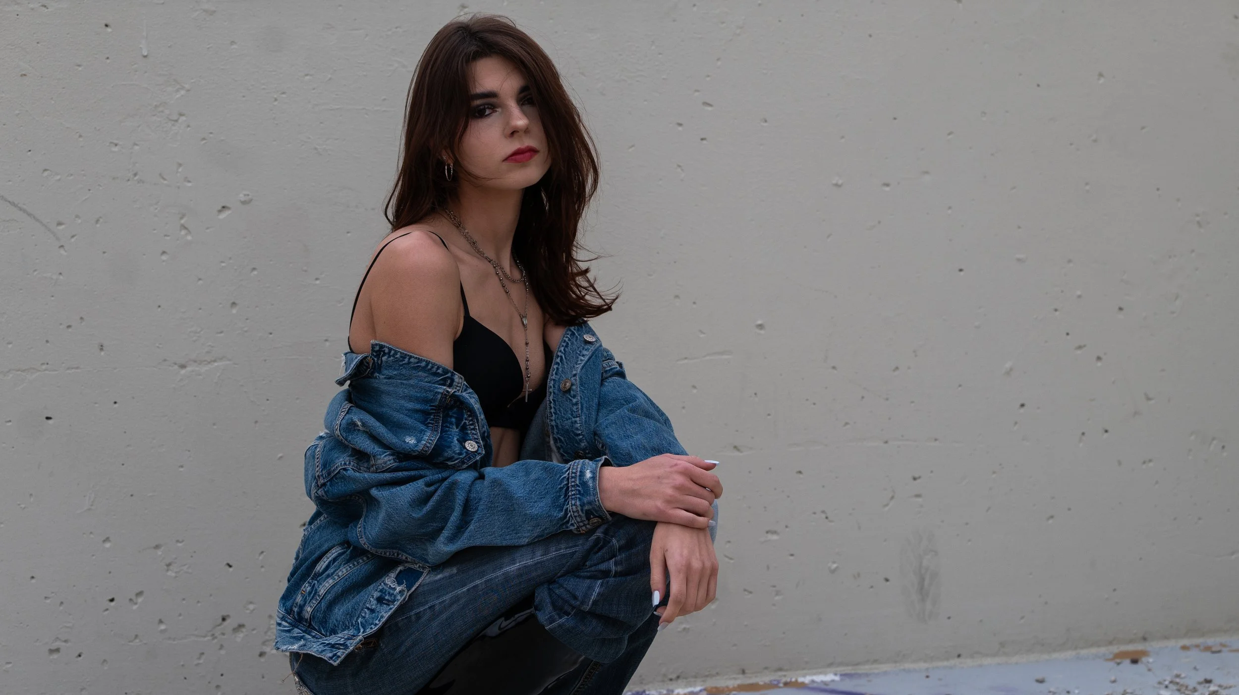 Young woman with dark hair wearing a black top and necklace, denim jacket off the shoulders, sitting against a textured gray wall.