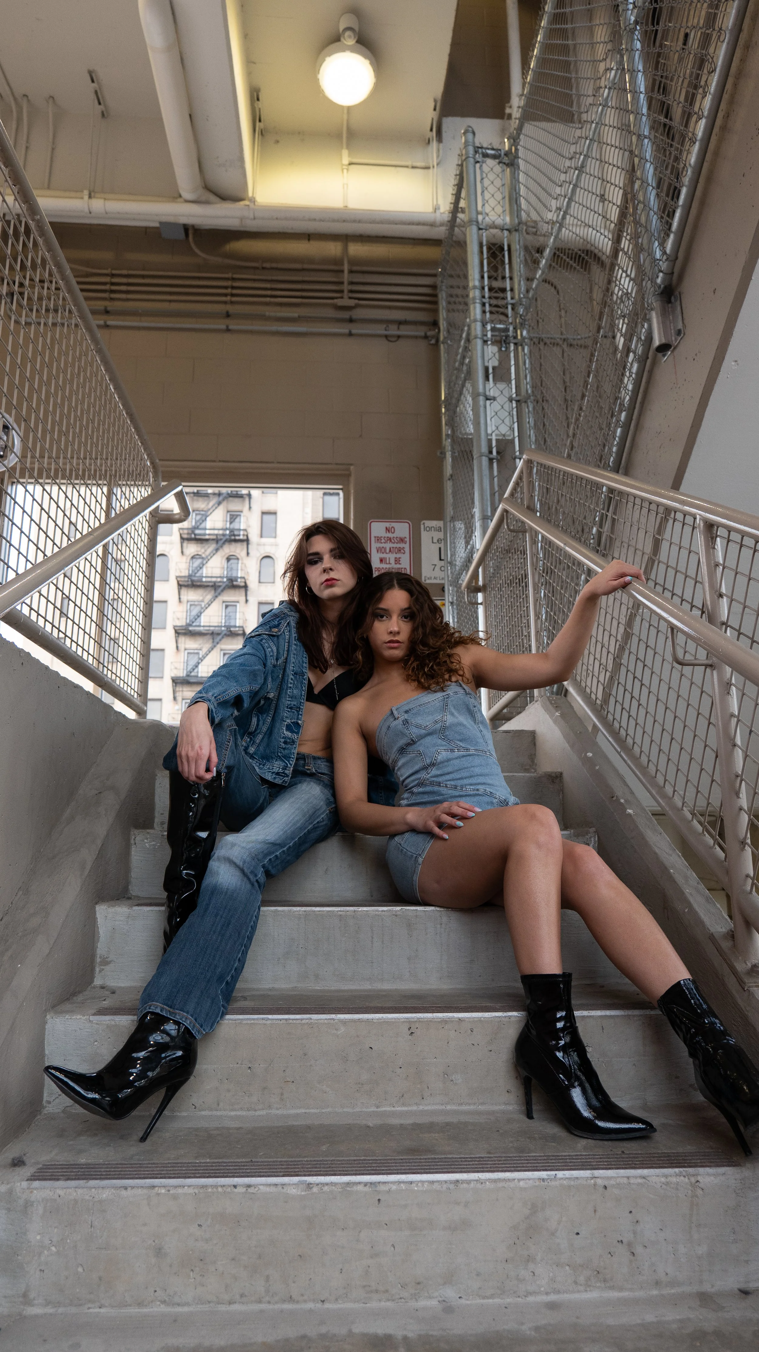 Two young women with dark brown and light brown curly hair posing on a concrete staircase in an urban setting. One is wearing a denim jacket, jeans, and black patent high-heeled boots, while the other is in a strapless denim dress with black patent h