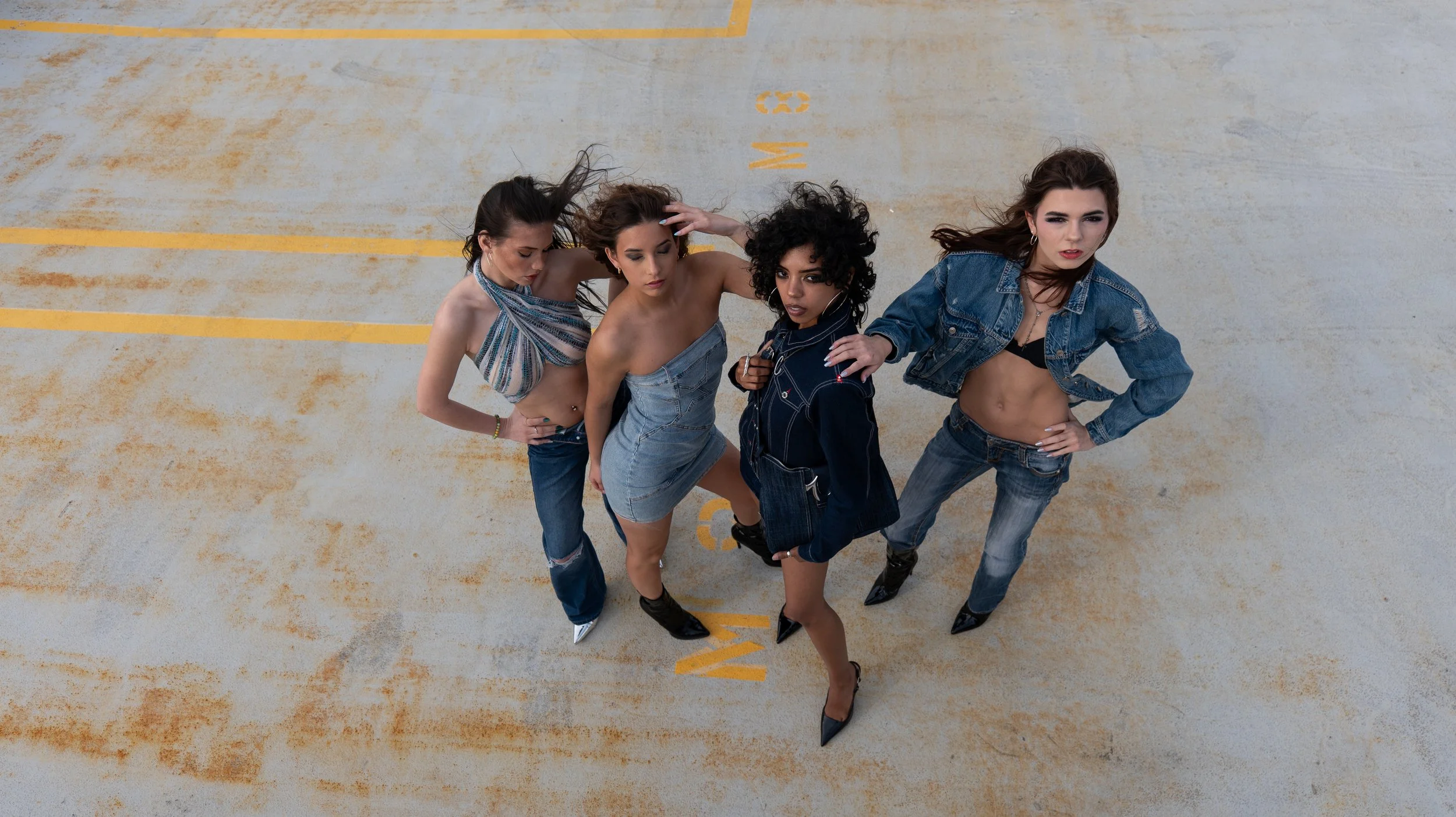 Four women standing together on a parking lot, looking up at the camera, with two of them wearing denim and two in tops with denim accents.
