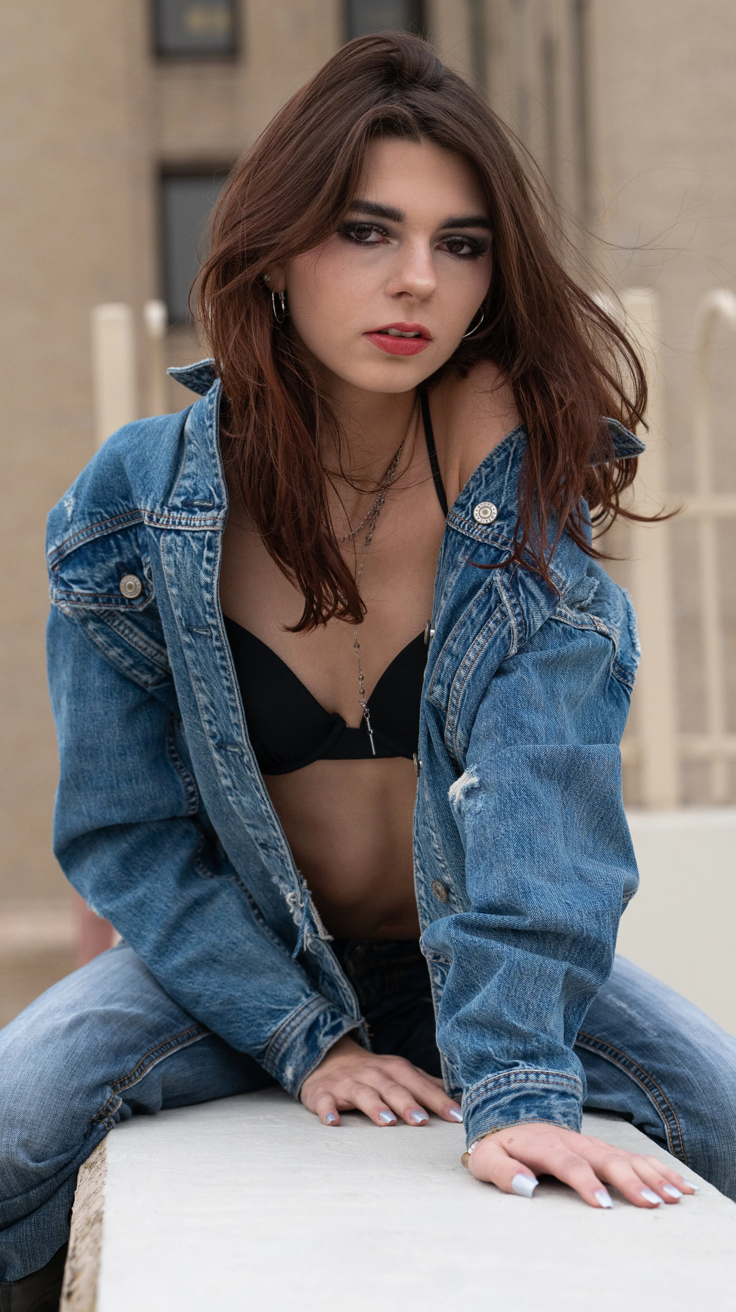 A young woman with brown hair and makeup, wearing a black bra and a blue denim jacket, sitting on the ground with one hand on a white surface and the other on her knee, looking into the camera.
