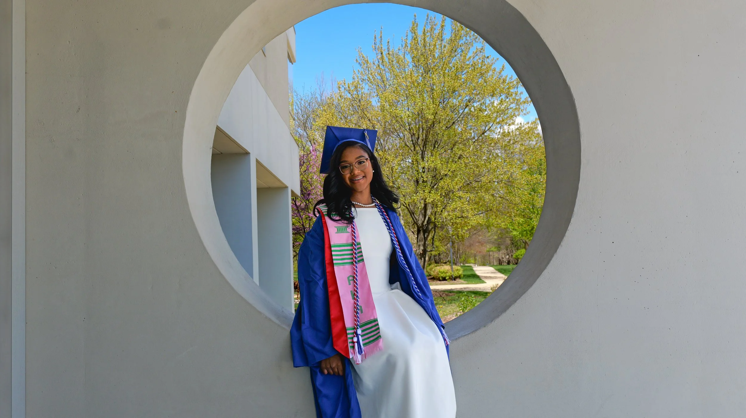A young woman in a graduation cap and gown smiling and posing outdoors, seen through a circular opening in a white wall, with trees and a sidewalk in the background.