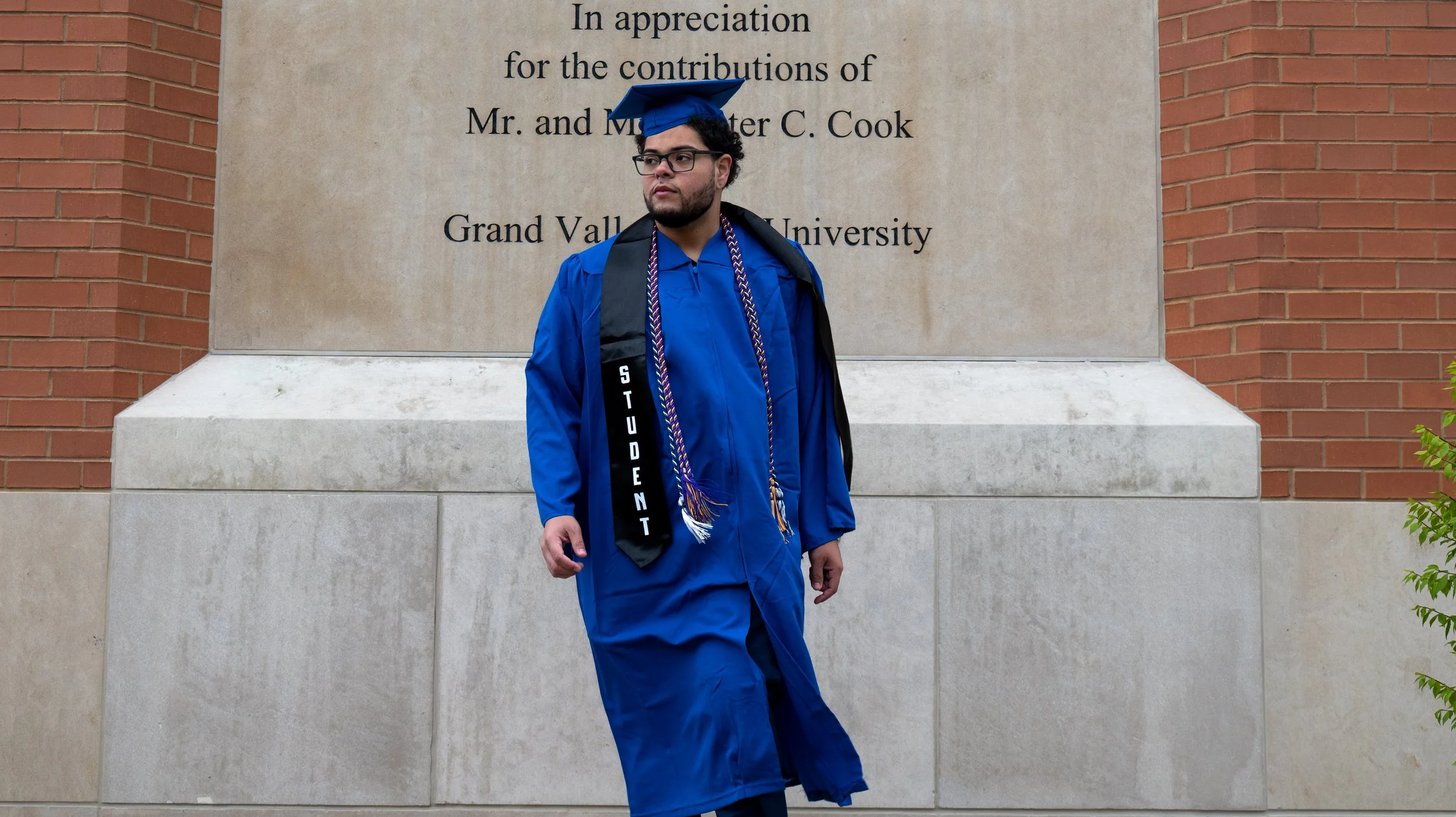 A young man in blue graduation gown and cap standing in front of a stone and brick wall with a plaque that has text about appreciation and contributions, at Grand Valley State University.