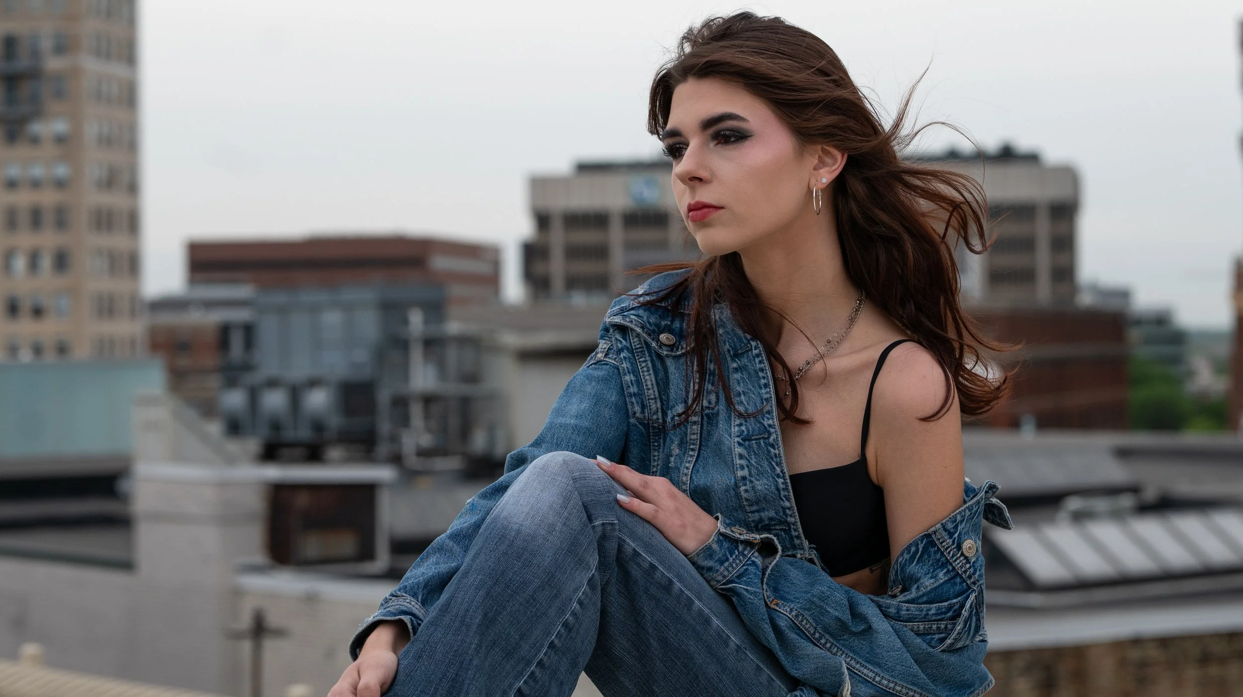 A young woman with brown hair, wearing a denim jacket and black top, sitting on a rooftop with city buildings in the background on an overcast day.