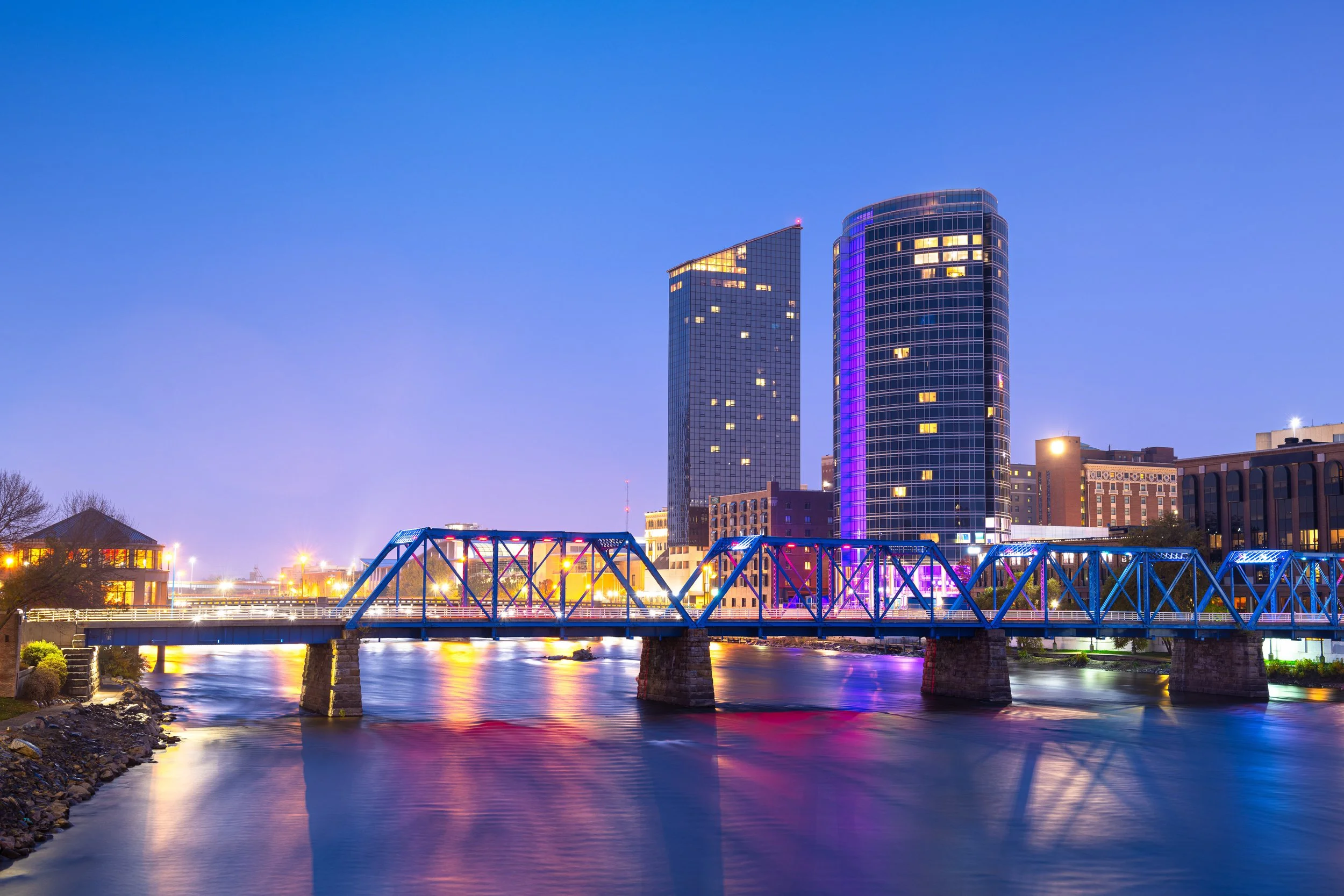 Night view of a city skyline featuring two tall modern glass buildings illuminated with colorful lights, a bridge over a river, and reflections on the water.