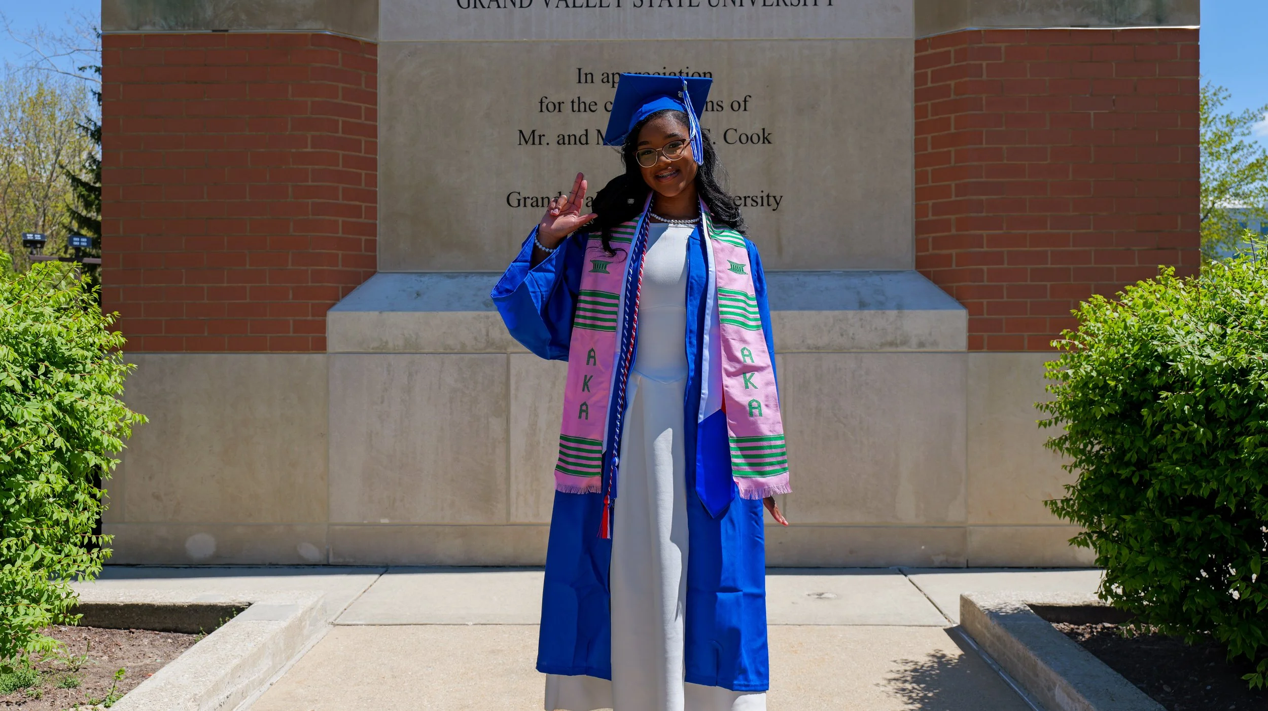 A young woman in a blue graduation gown and cap, smiling and making a peace sign while standing in front of a university monument with greenery on both sides.