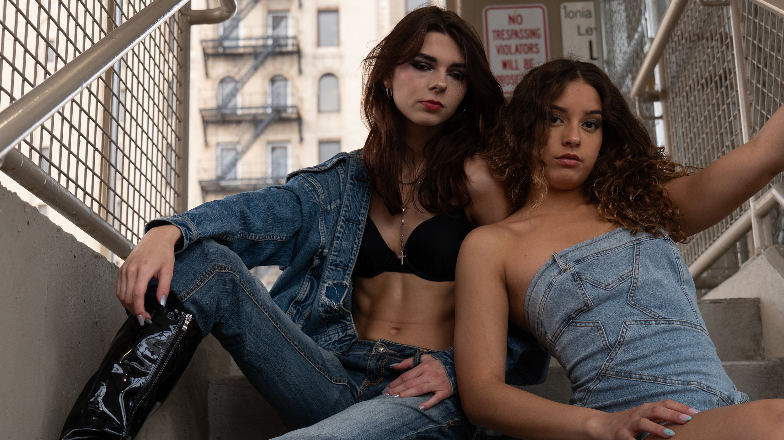 Two young women sitting on concrete stairs outdoors, surrounded by metal railings. One woman has brown hair, wears a denim jacket and jeans, and holds a camera; the other has curly hair, wears a denim jacket, and is topless. Background includes urban