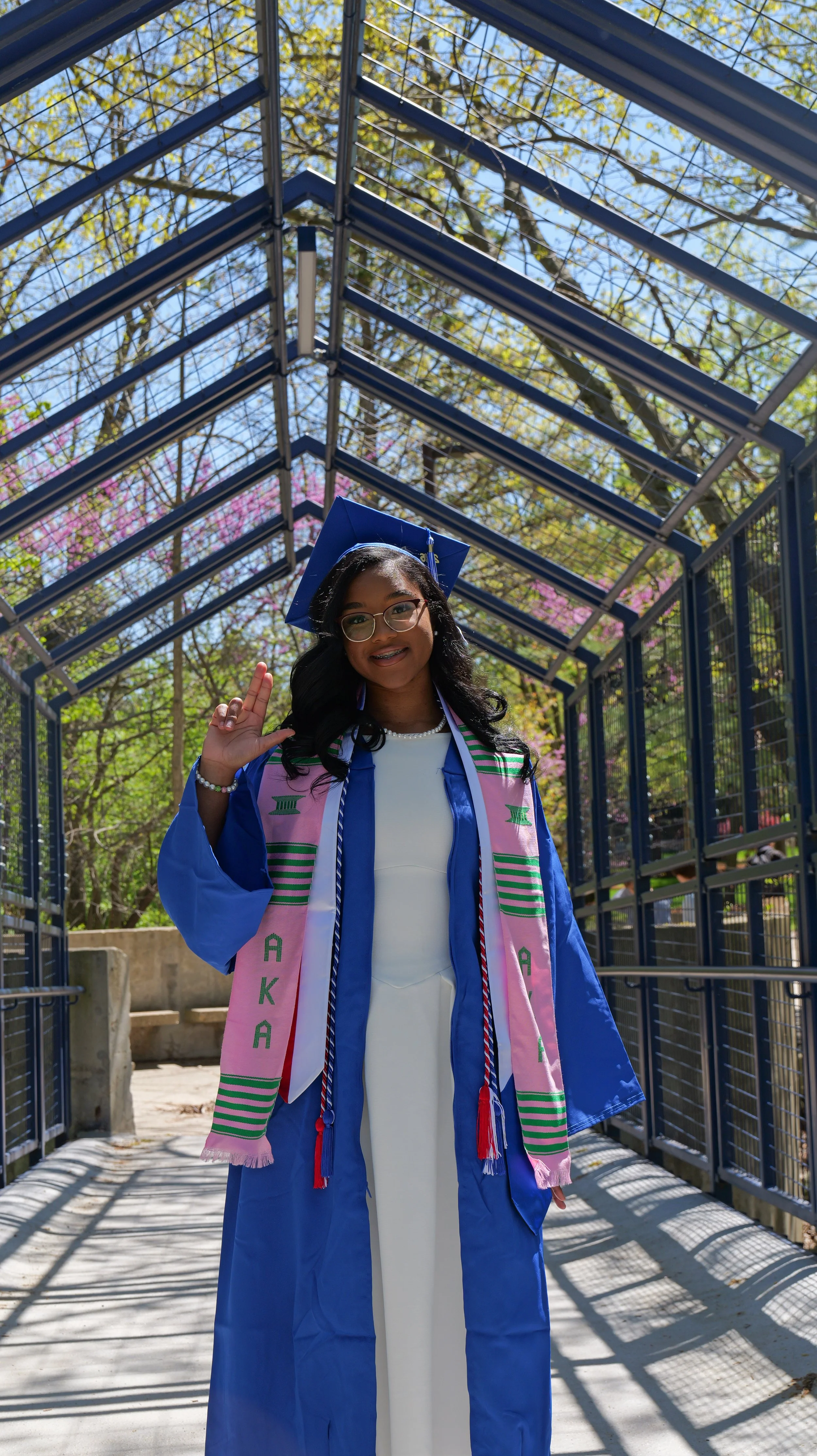 Young woman in a blue graduation gown and cap standing outdoors under a metal structure with trees in the background. She is smiling, making a hand gesture, and is wearing a pink stole with green and white accents.
