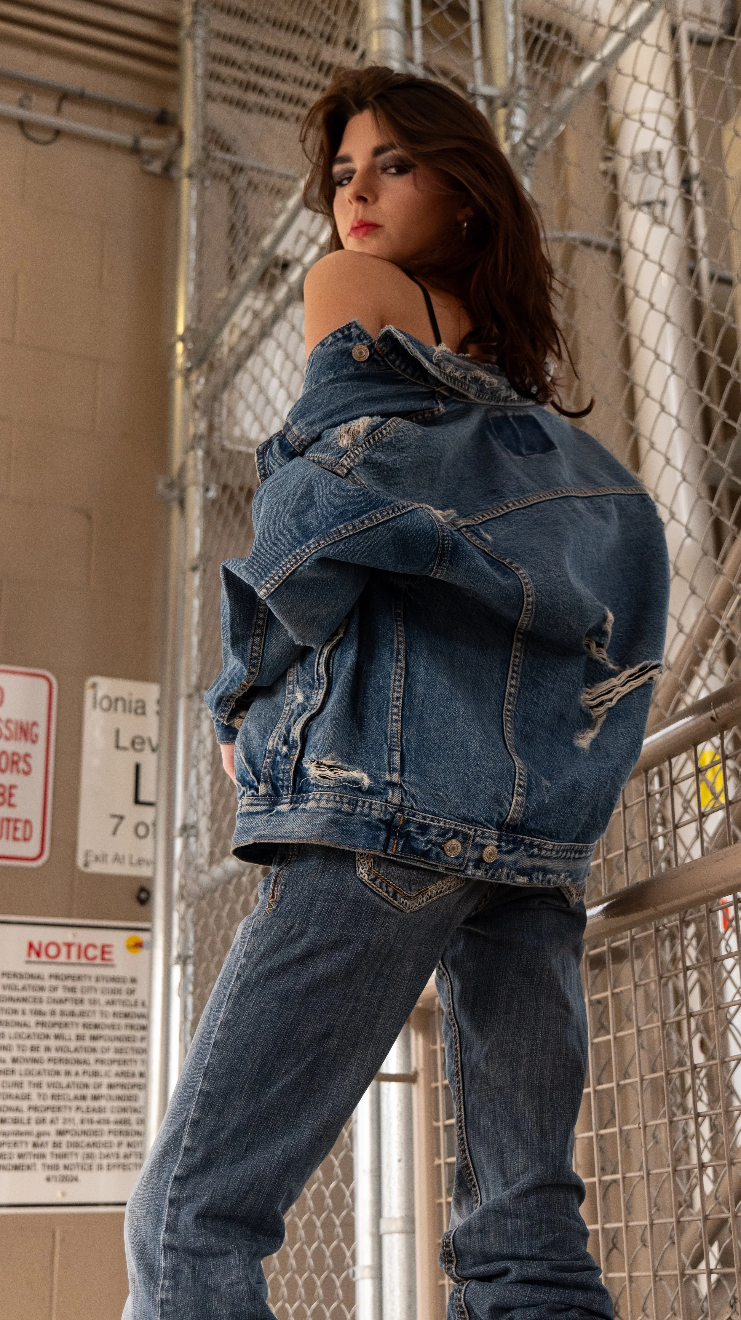 Young woman with brown hair wearing a ripped jean jacket and jeans, standing against a chain-link fence in an industrial setting.