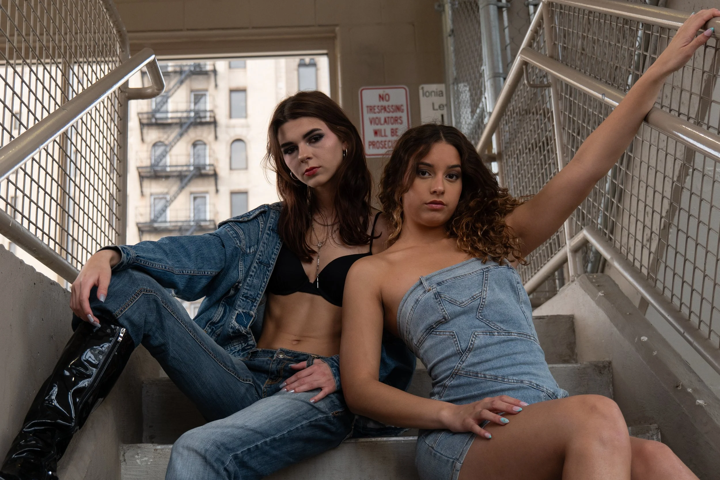 Two young women sitting on outdoor stairs, one wearing a denim jacket and jeans, the other in a black top and distressed jeans, with urban building background.
