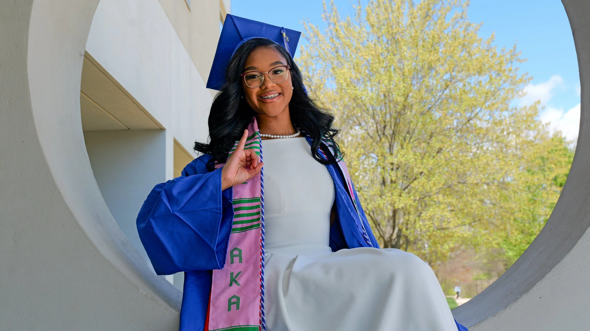 Young woman in graduation cap and gown sitting outdoors near a large circular opening, smiling, with trees and blue sky in the background.