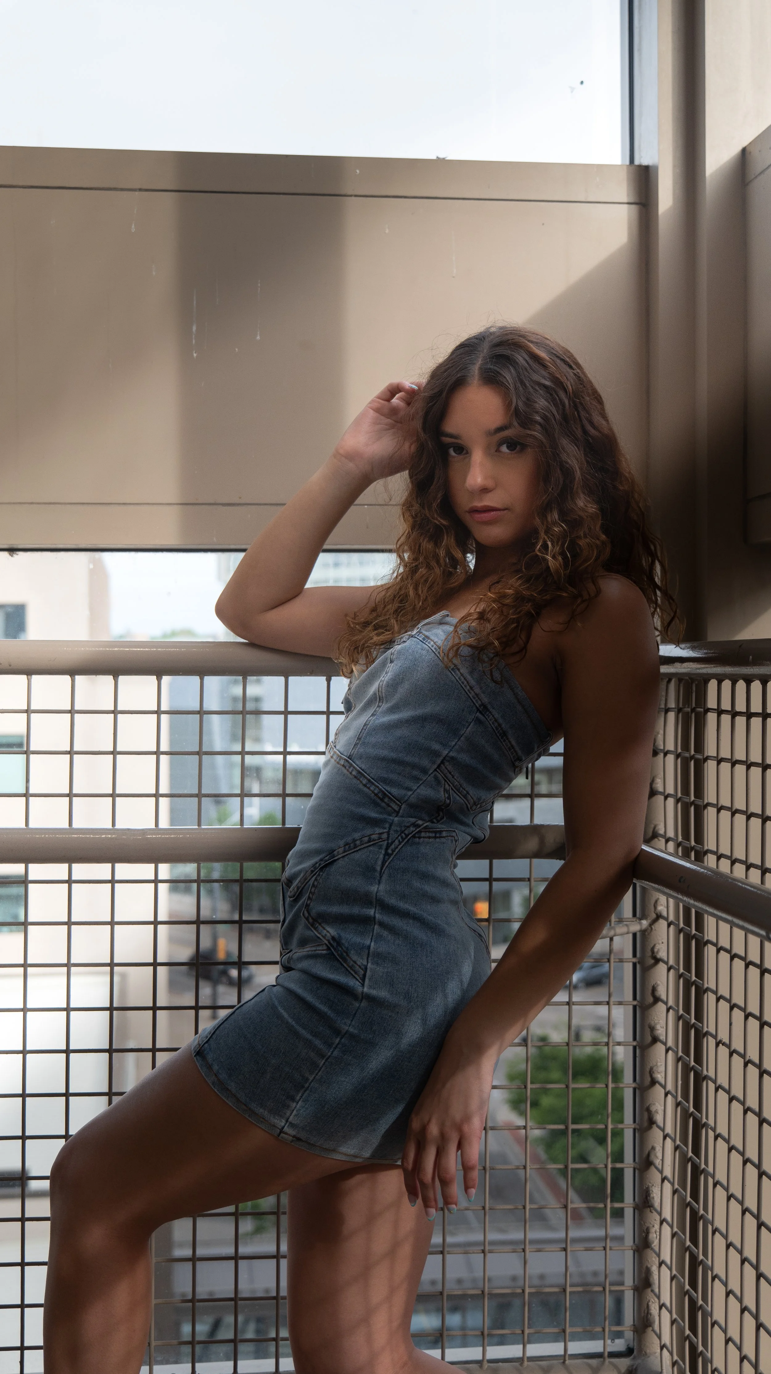 A woman in denim overalls leaning against a metal railing on a balcony with city buildings and trees in the background.