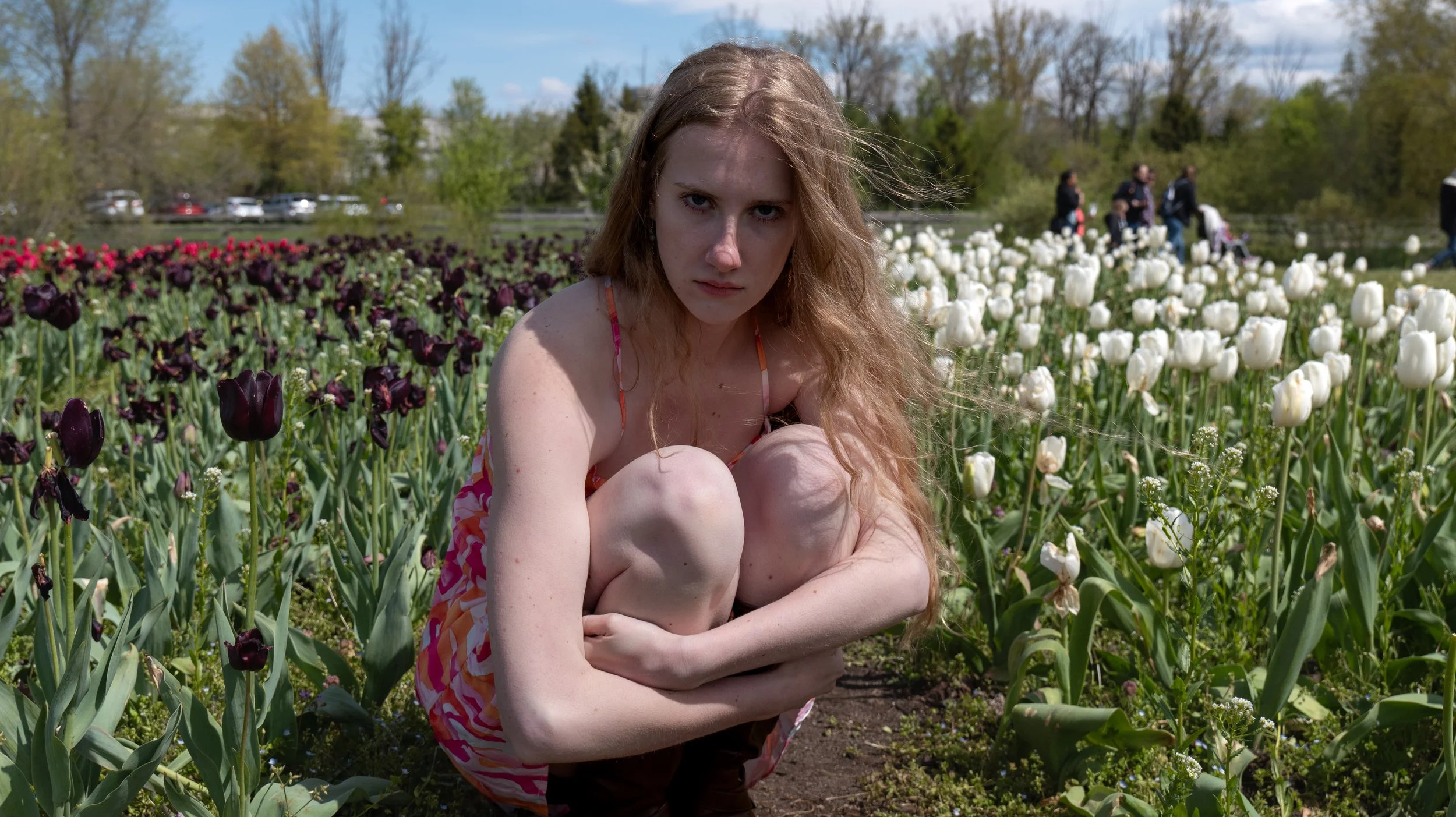 Young woman with long red hair in a colorful dress sitting among tulips in a flower garden on a sunny day.