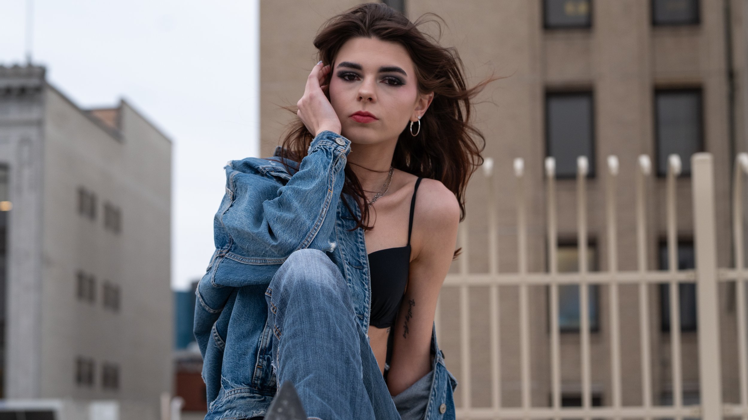 A woman with dark hair and makeup sitting outdoors with a city building in the background.