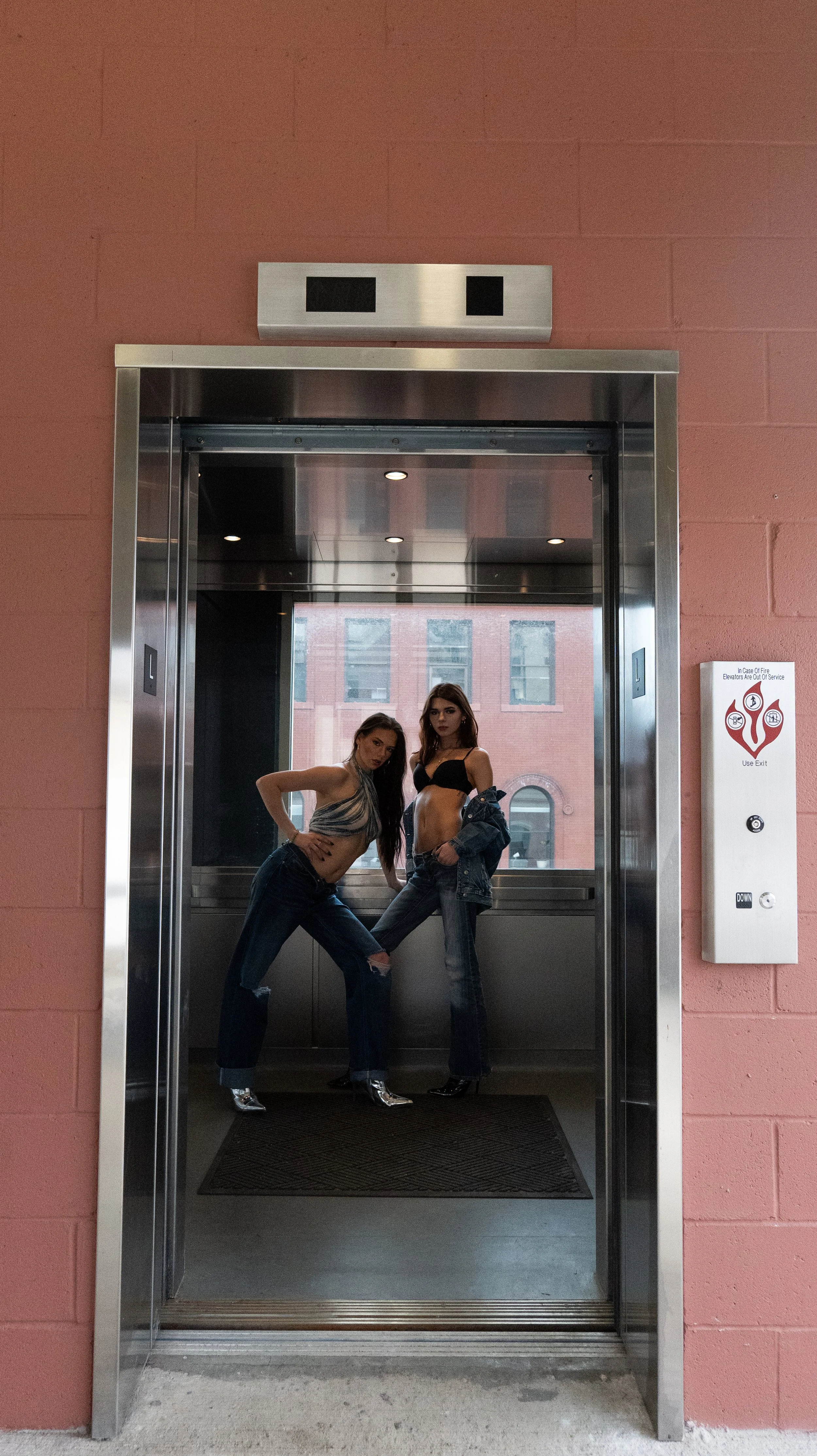 Two women with long hair and ripped jeans posing inside an elevator, wearing black tops and heeled shoes, with a city building visible through the windows.