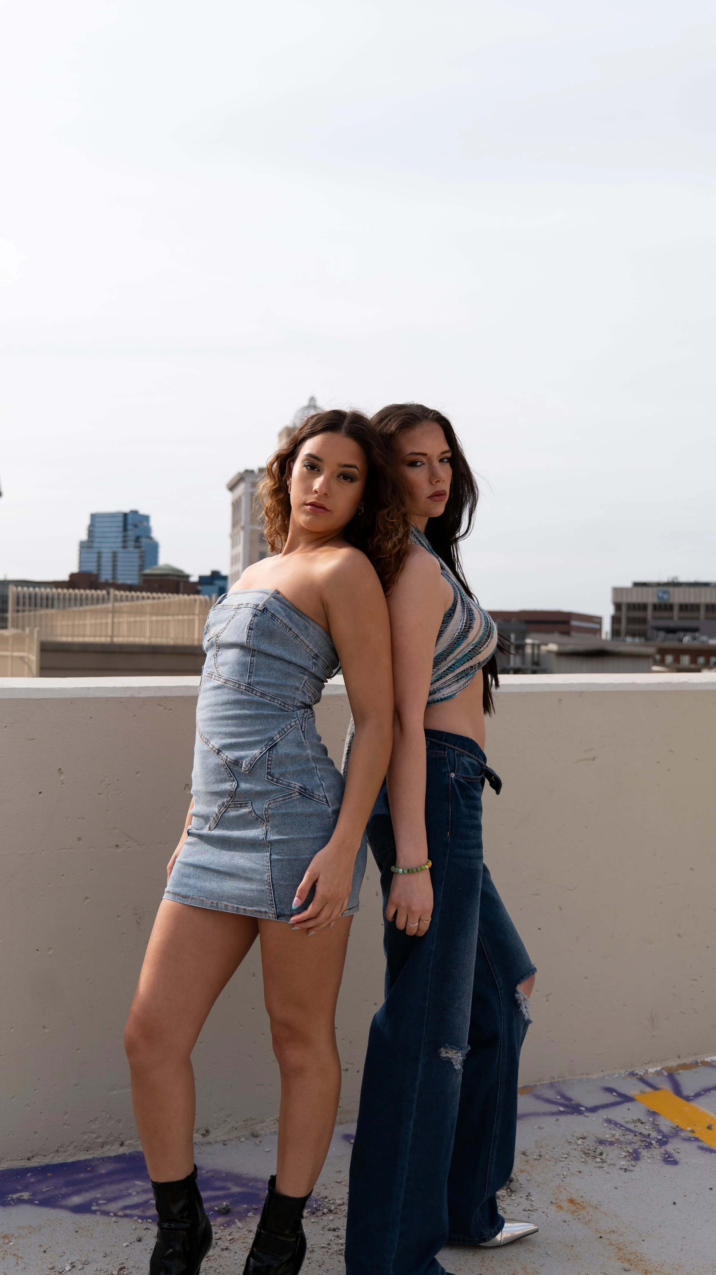 Two young women stand back-to-back on a rooftop, wearing casual denim outfits, with a city skyline in the background under a cloudy sky.