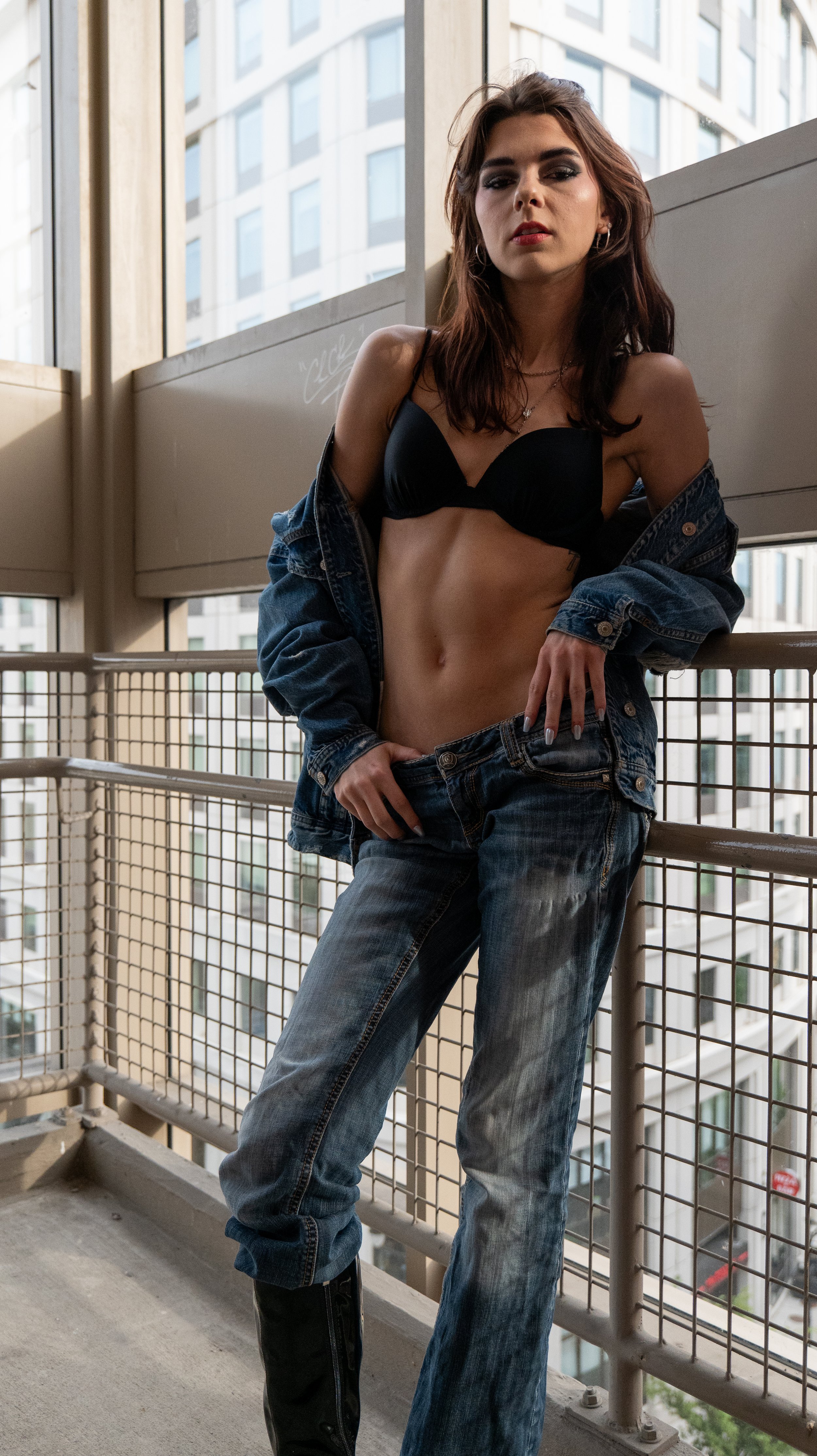 Young woman with brown hair and makeup, wearing a black bra, denim jacket, and jeans, posed on a balcony with city buildings in the background.