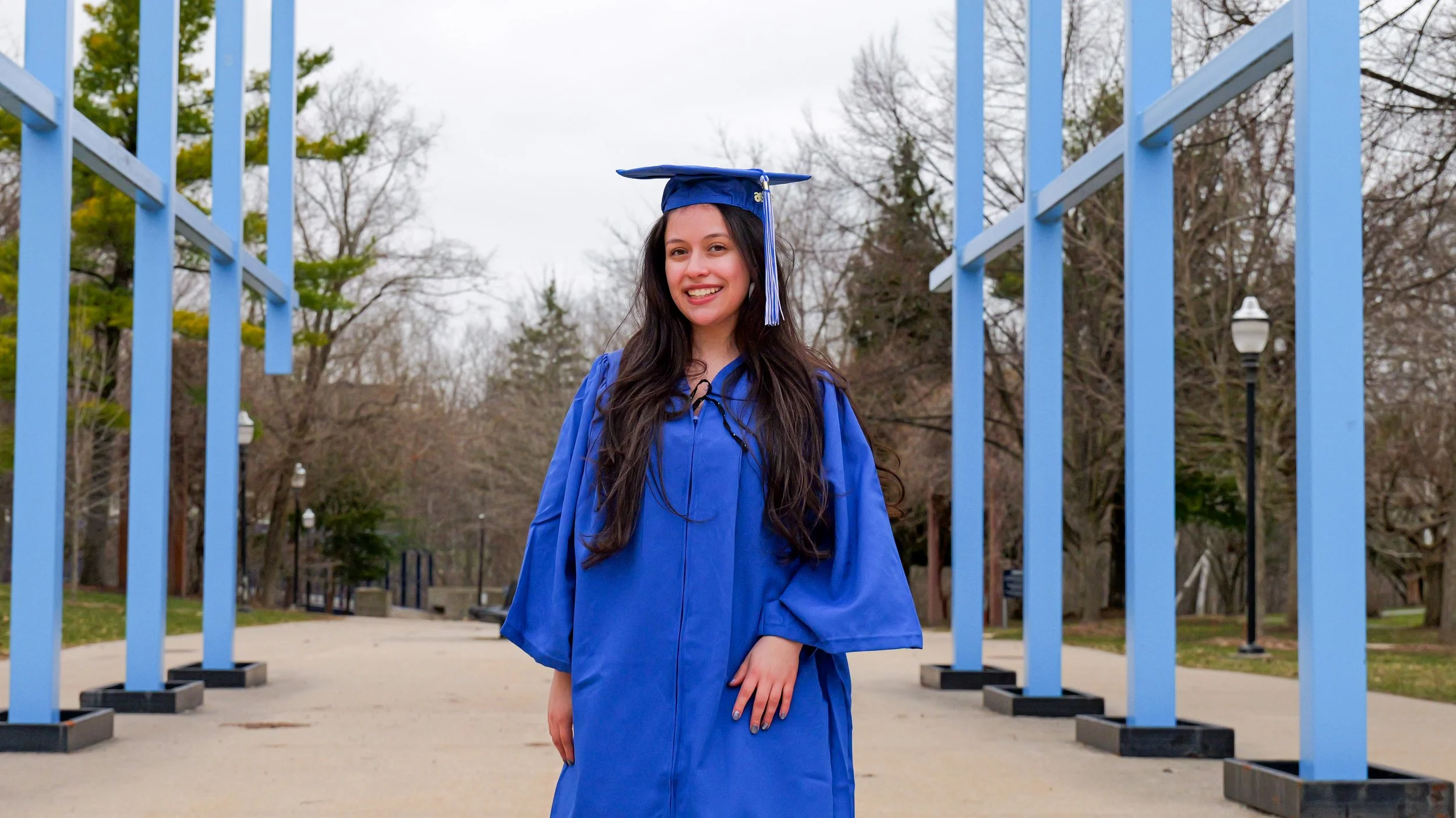 A young woman in a blue graduation cap and gown standing outdoors, smiling, with blue decorative structures and trees in the background.