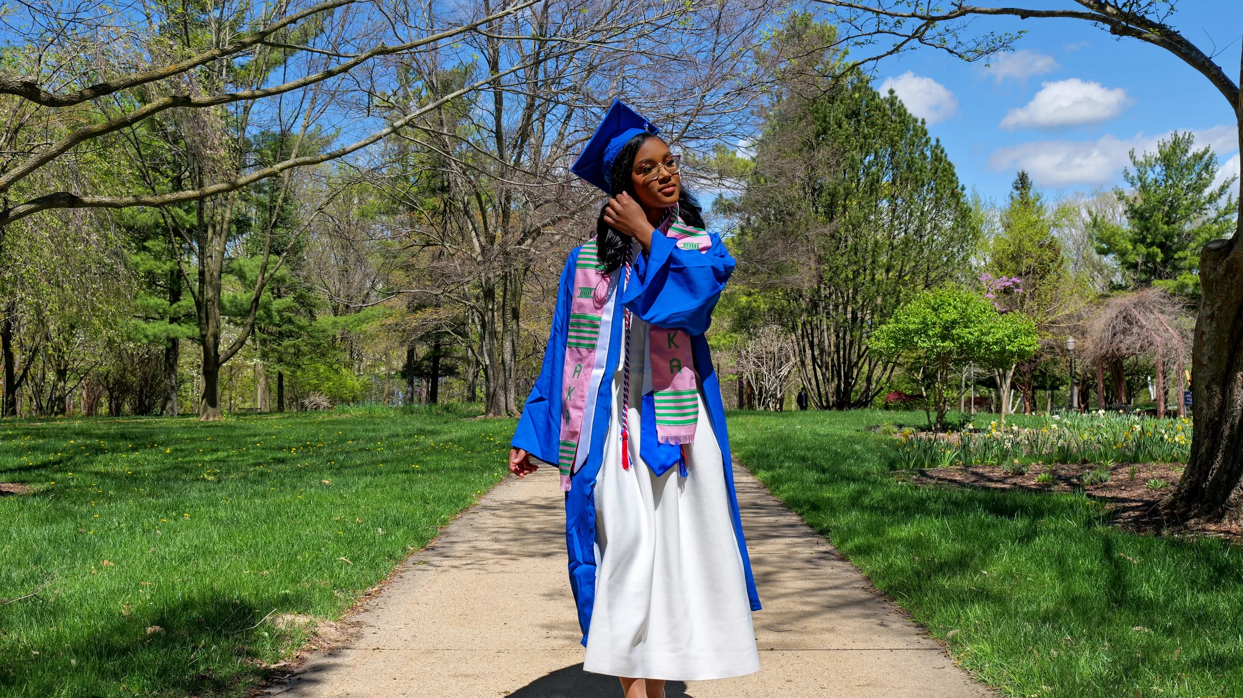 A woman in a blue graduation gown and cap standing on a park path with trees and green grass.
