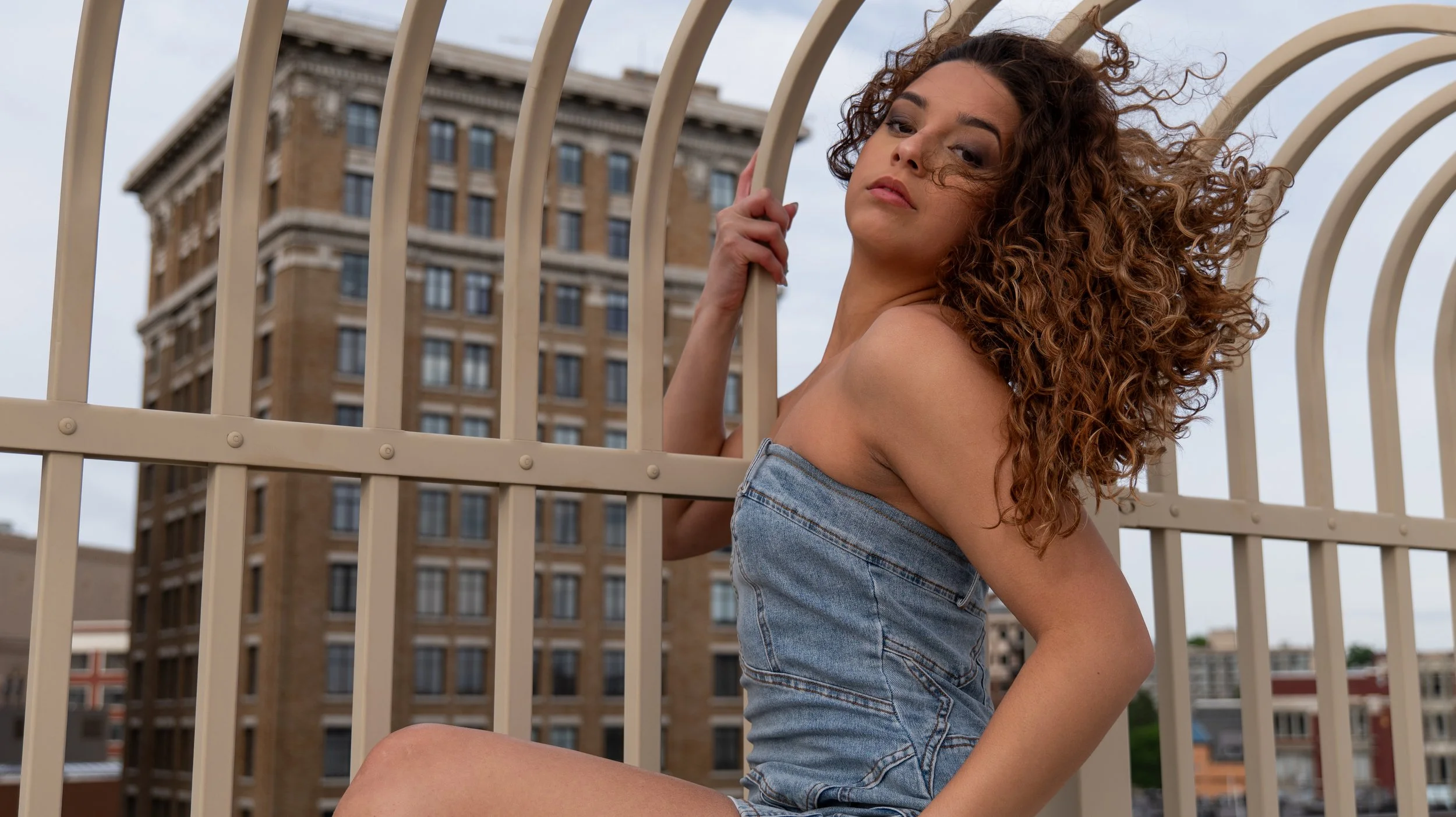A woman with curly hair leaning against a beige metal railing on a rooftop, with a city building in the background under an overcast sky.