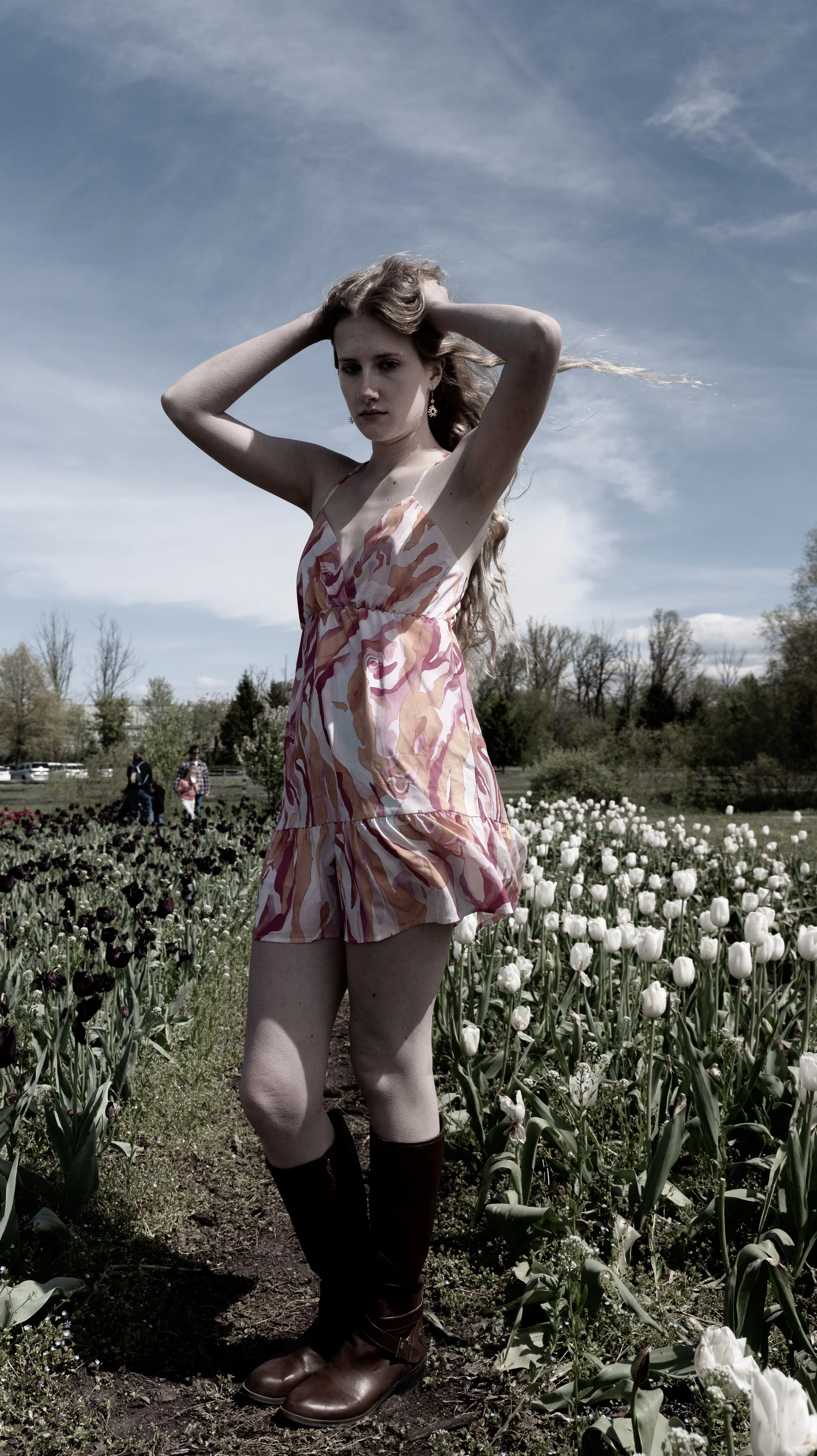 Young woman in a patterned dress and tall boots standing in a tulip field under a partly cloudy sky.