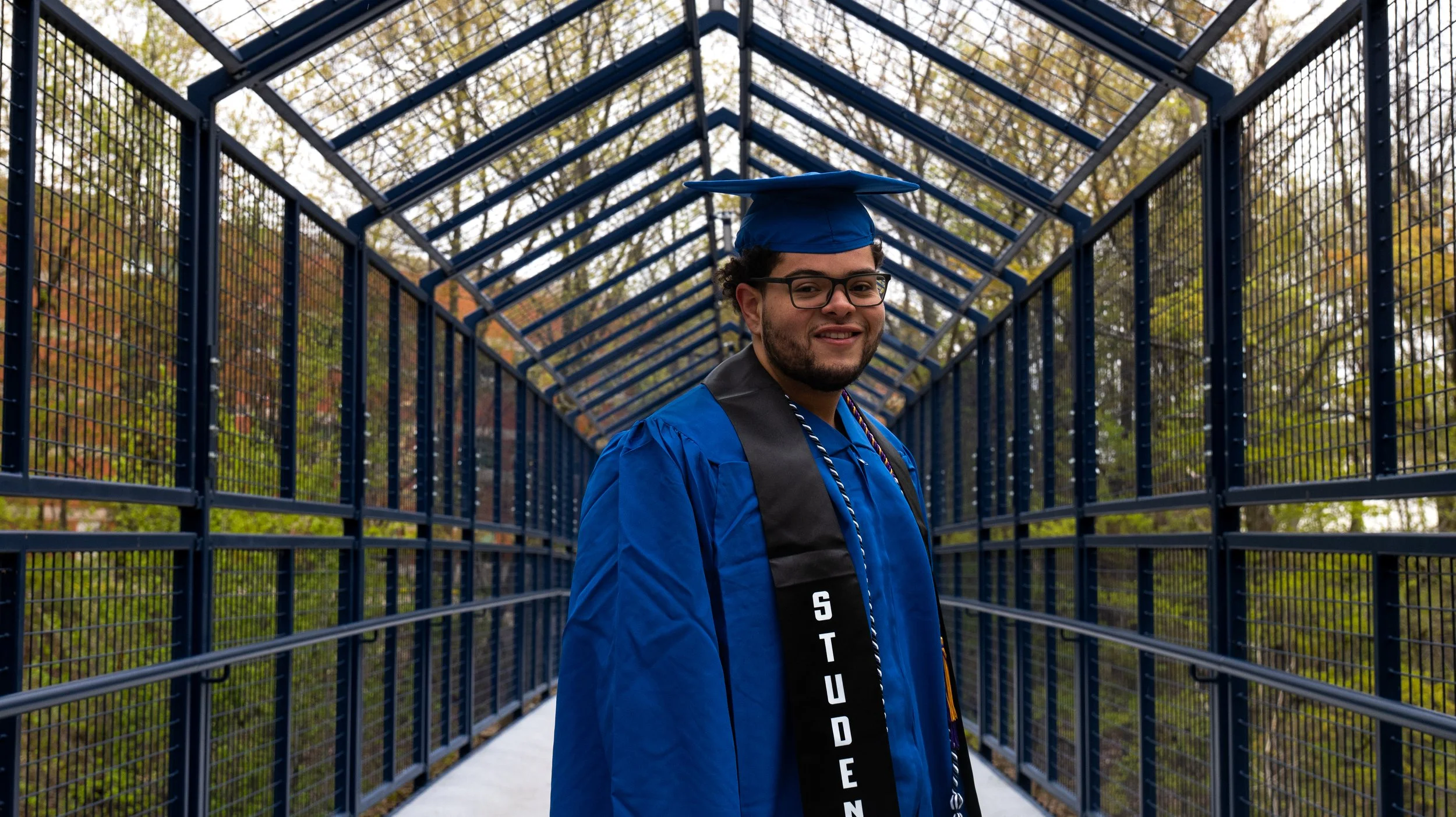 A young man in a blue graduation cap and gown standing in the middle of an outdoor blue metal walkway with trees in the background.