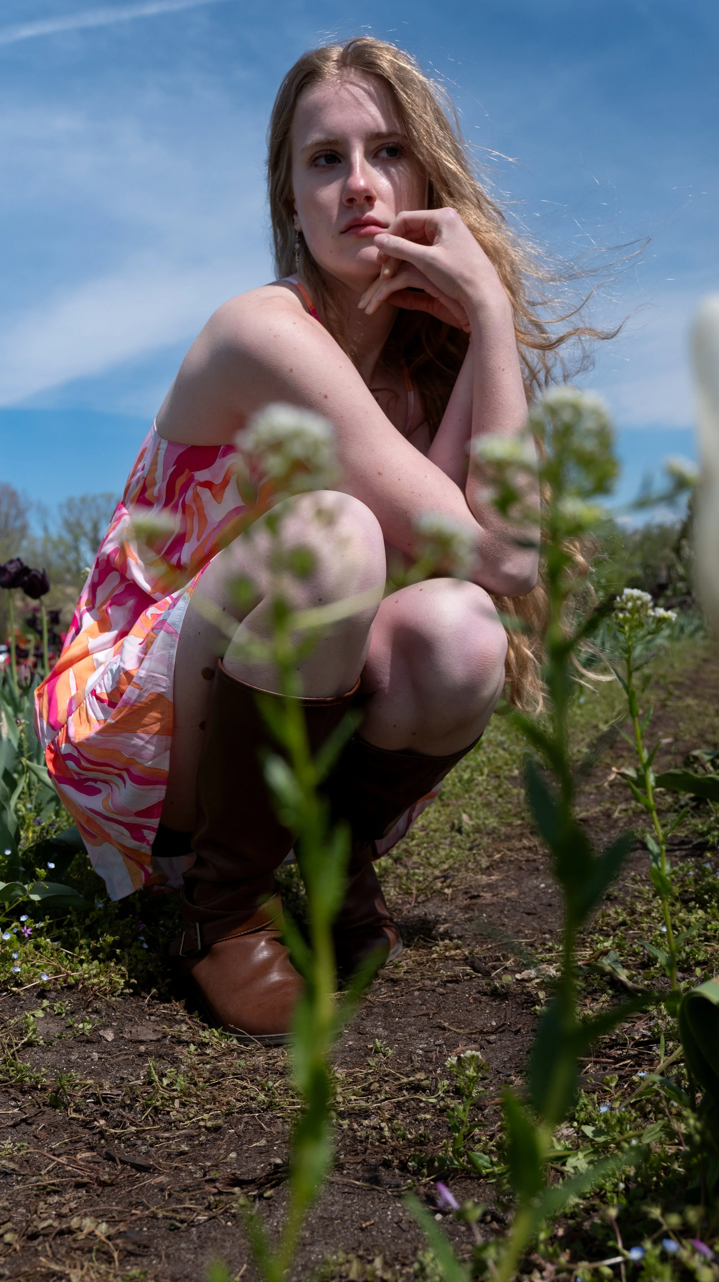 Young woman with long red hair in a floral dress squatting in a flower field with a clear blue sky overhead.