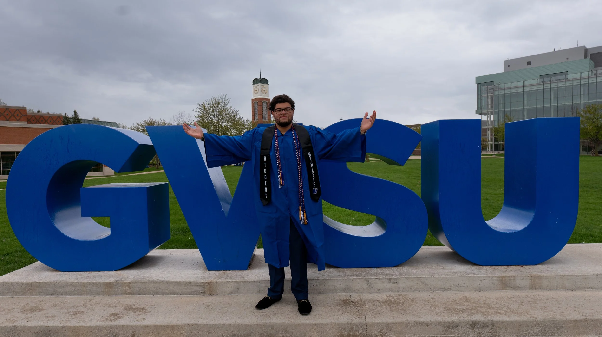 A young man in a blue graduation cap and gown with honor cords stands with arms outstretched in front of large blue letters spelling 'GVSU' on a university campus with a clock tower, brick buildings, and a cloudy sky in the background.