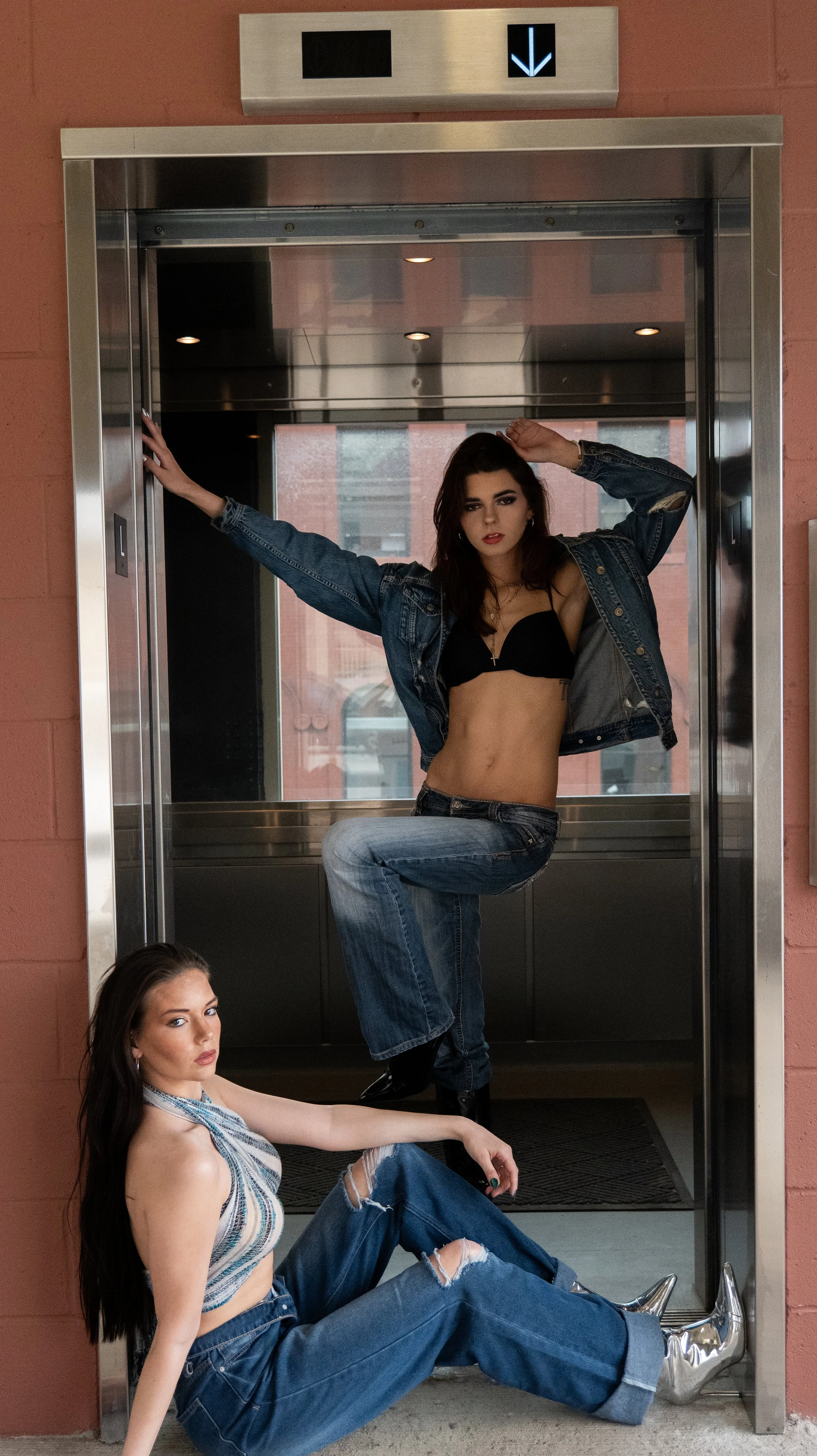 Three women in casual clothing posing in an elevator and on the floor in front of it, with a black and blue downward arrow sign above.