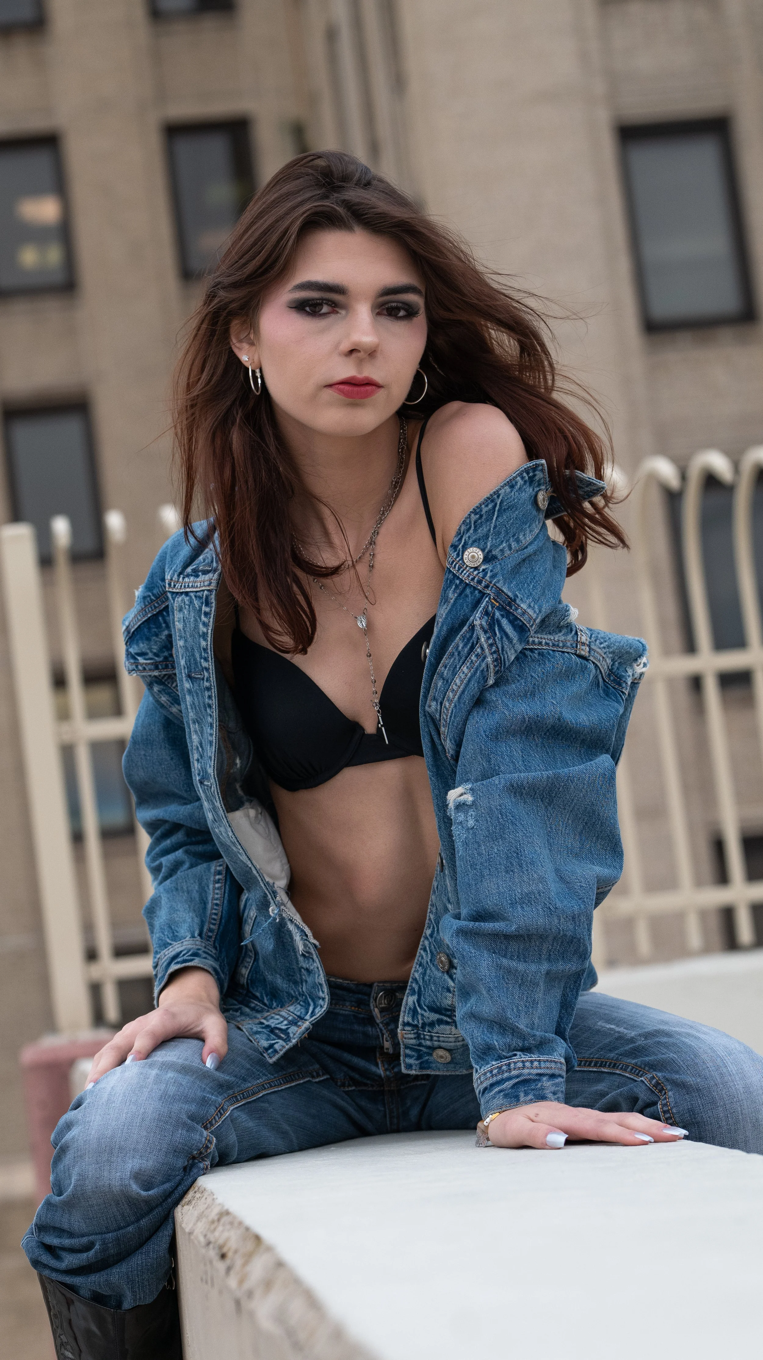 Young woman with brown hair, wearing a black bra, a denim jacket, and jeans, sitting outdoors on a concrete surface, looking at the camera with a serious expression.