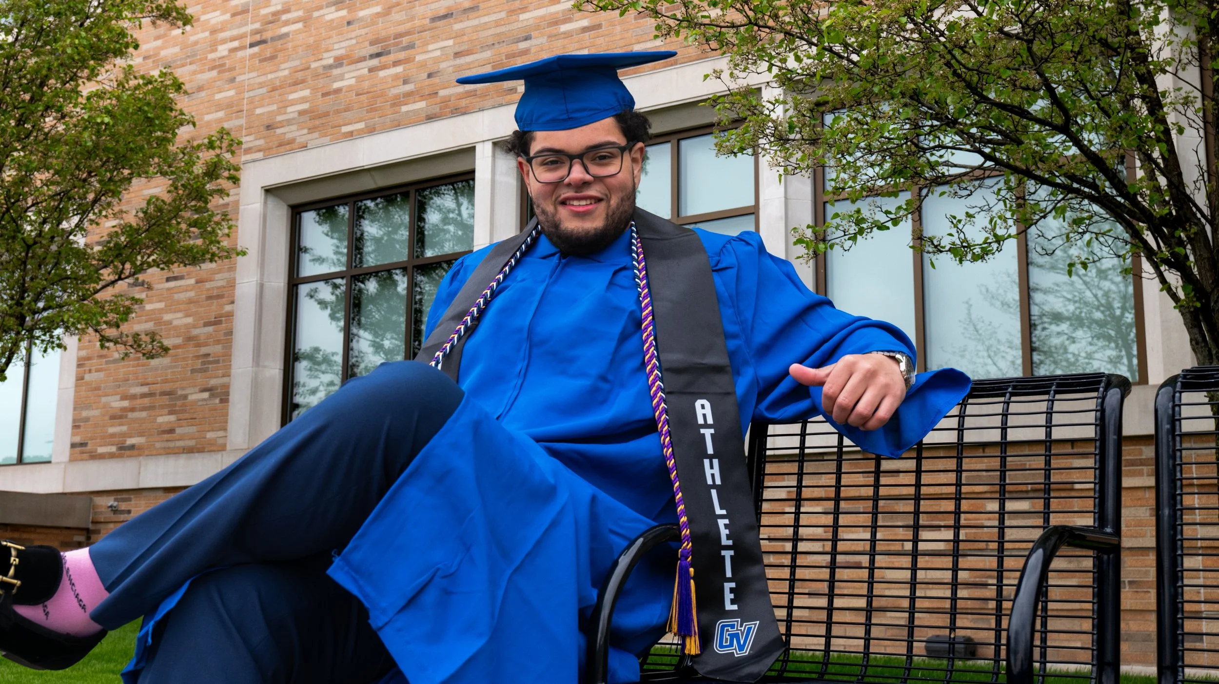 Young man in blue graduation cap and gown sitting on a bench outdoors in front of a brick building with large windows, smiling at the camera.