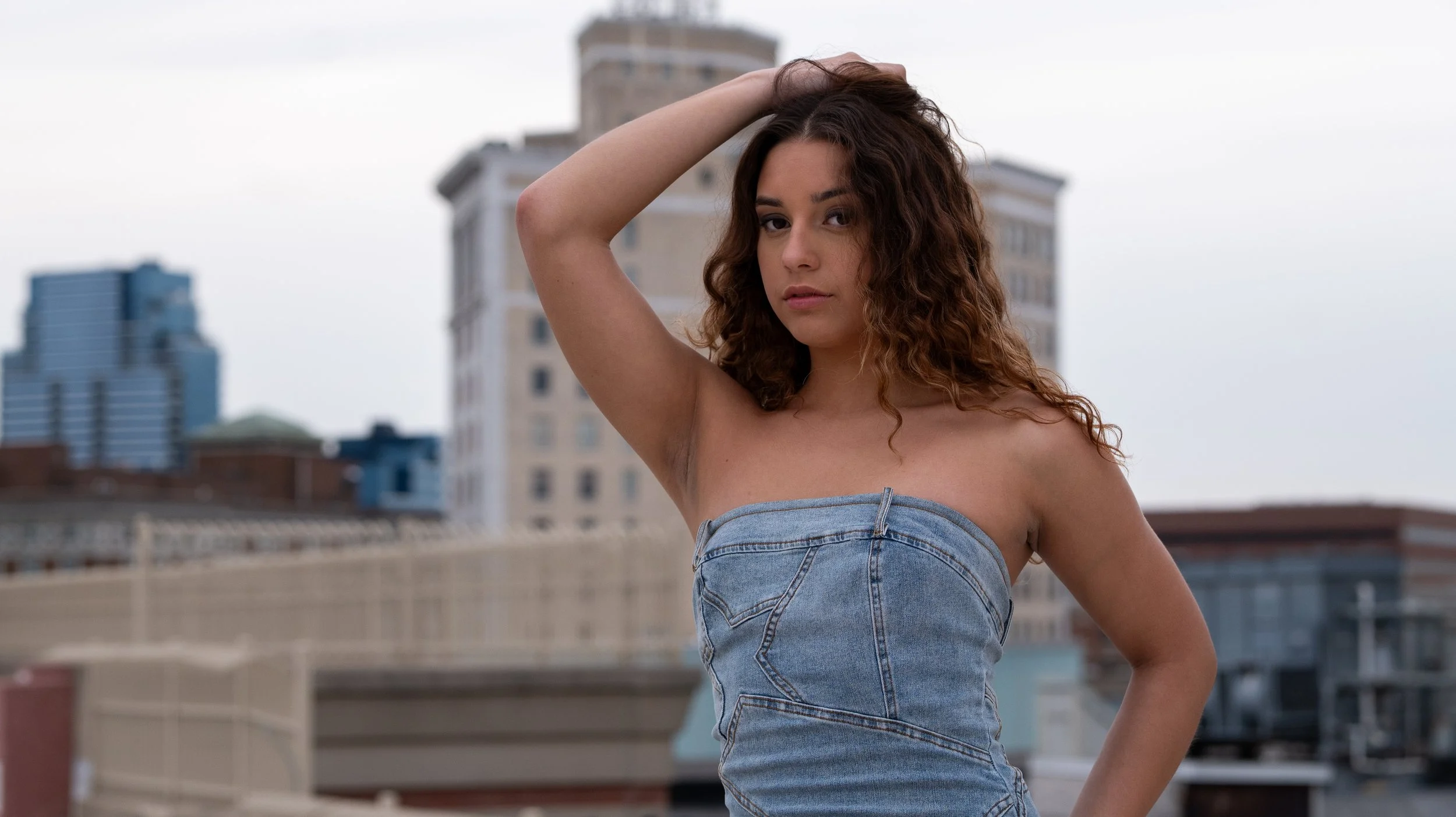 A young woman with curly brown hair wearing a strapless denim top standing on a rooftop with city buildings in the background.