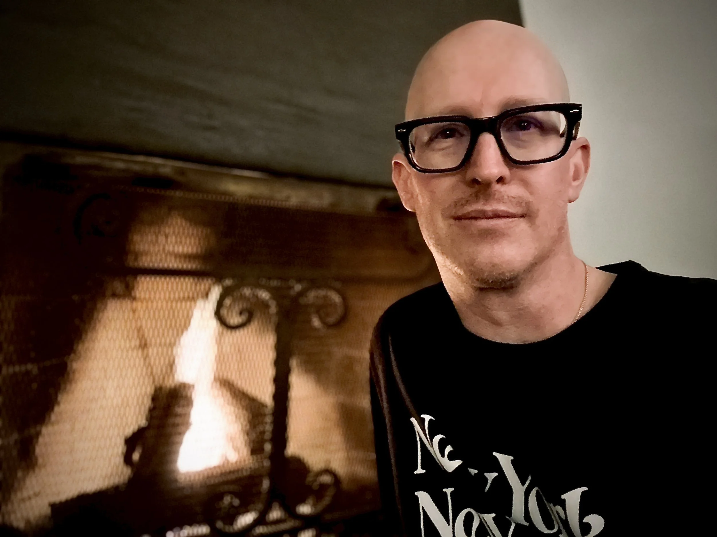 A person with a bald head, black glasses, and a slight smile, sitting indoors in front of a fire place inviting you to joing a nice conversation.