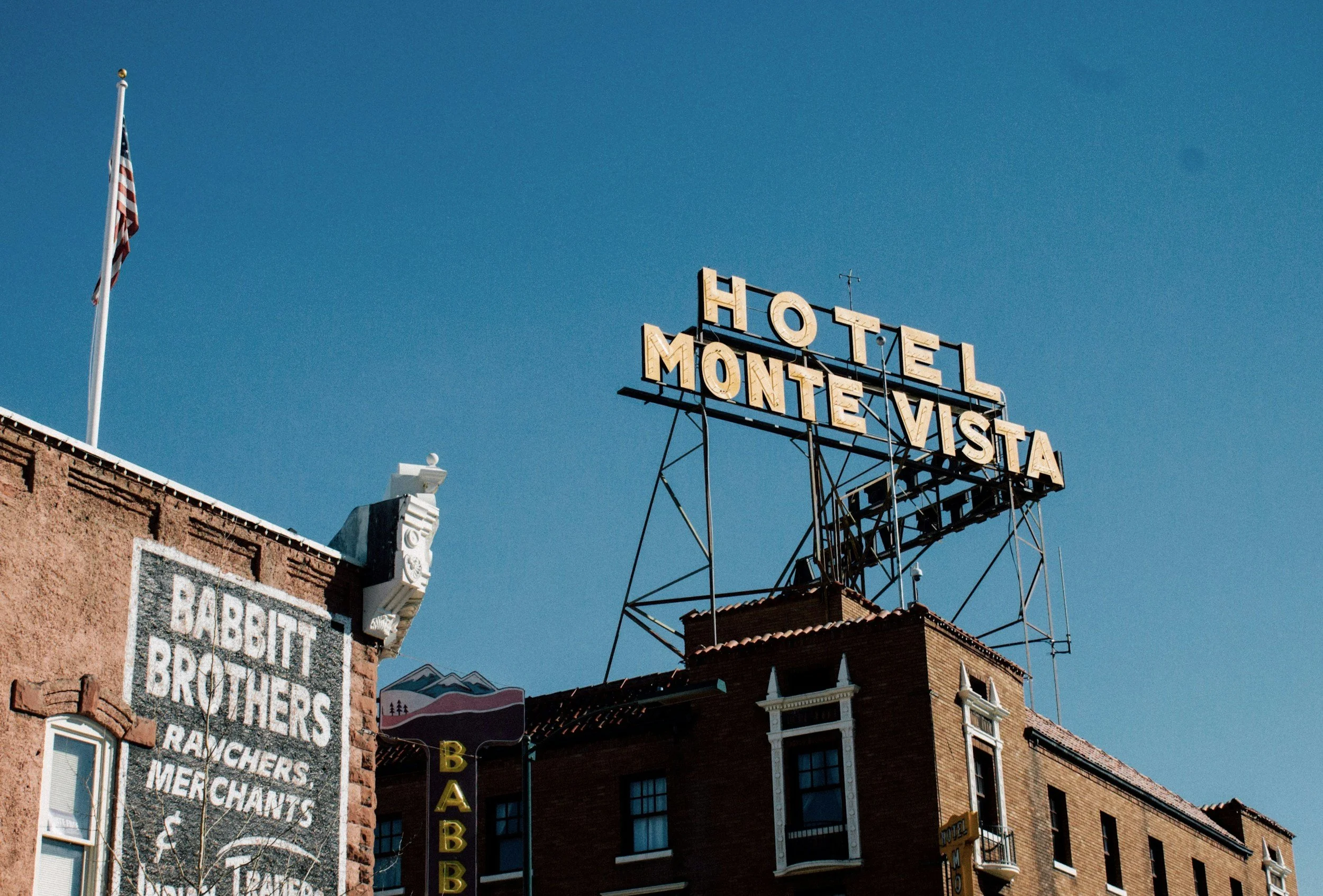 The image shows a blue sky with a vintage neon hotel sign reading 'HOTEL MONTE VISTA' mounted on a metal framework on top of a building. There is an American flag on a flagpole and part of a brick building with various signs, including a blackboard with service advertisements for rabbit brothers, ranchers, and merchandisers.