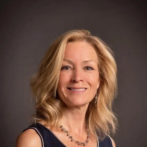 Headshot of a smiling woman with blonde, wavy hair, wearing earrings and a dark sleeveless top against a dark background.
