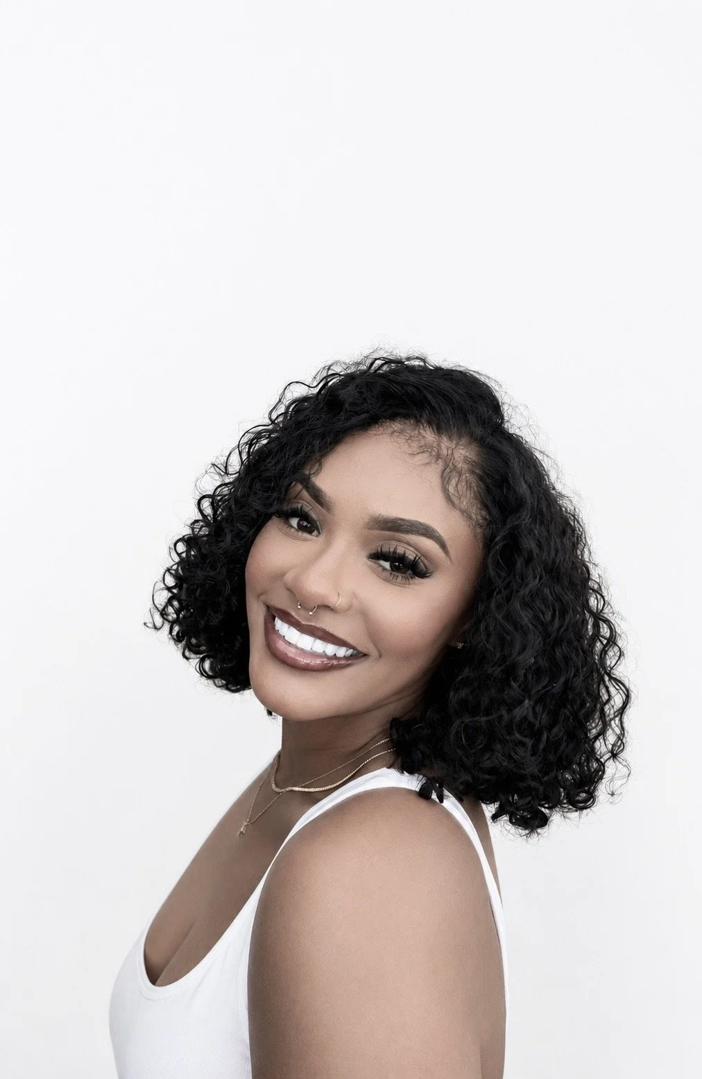 Portrait of a smiling woman with curly black hair, wearing a white sleeveless top and jewelry, posing against a white background.