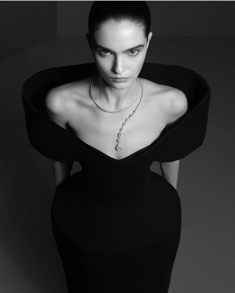 A black and white photograph of a woman with dark hair tied back, wearing an elegant black off-the-shoulder dress and a diamond necklace, looking directly at the camera with an intense expression.