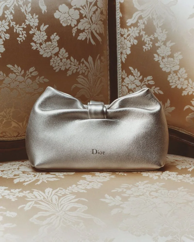 A metallic silver Dior purse with a large bow on top, placed against an ornate floral patterned background and surface.